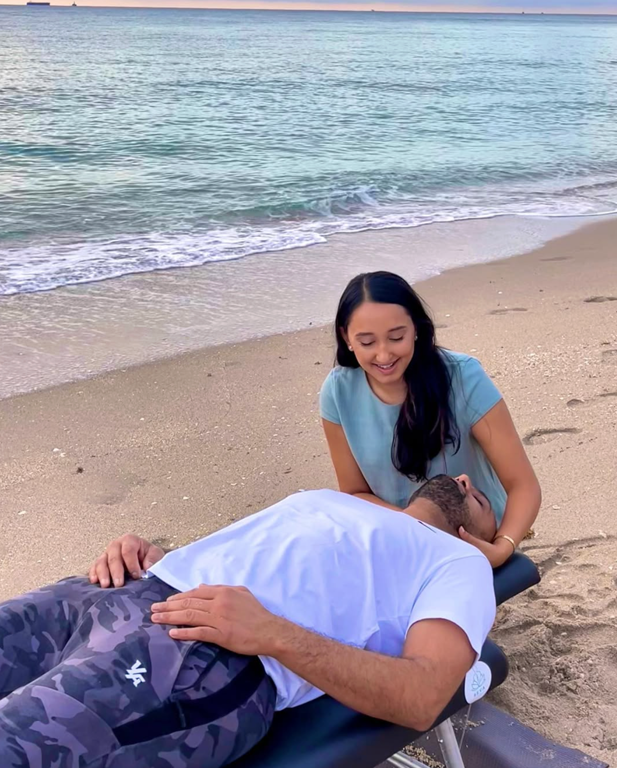Two people on a beach, one lying down while the other kneels beside them near the shoreline.