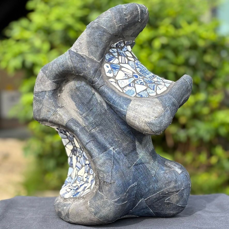 Een abstract kunstwerk, gemaakt van getextureerde, donkerblauwe steen met accenten van witte en blauwe mozaïektegels, geplaatst in de buitenlucht.