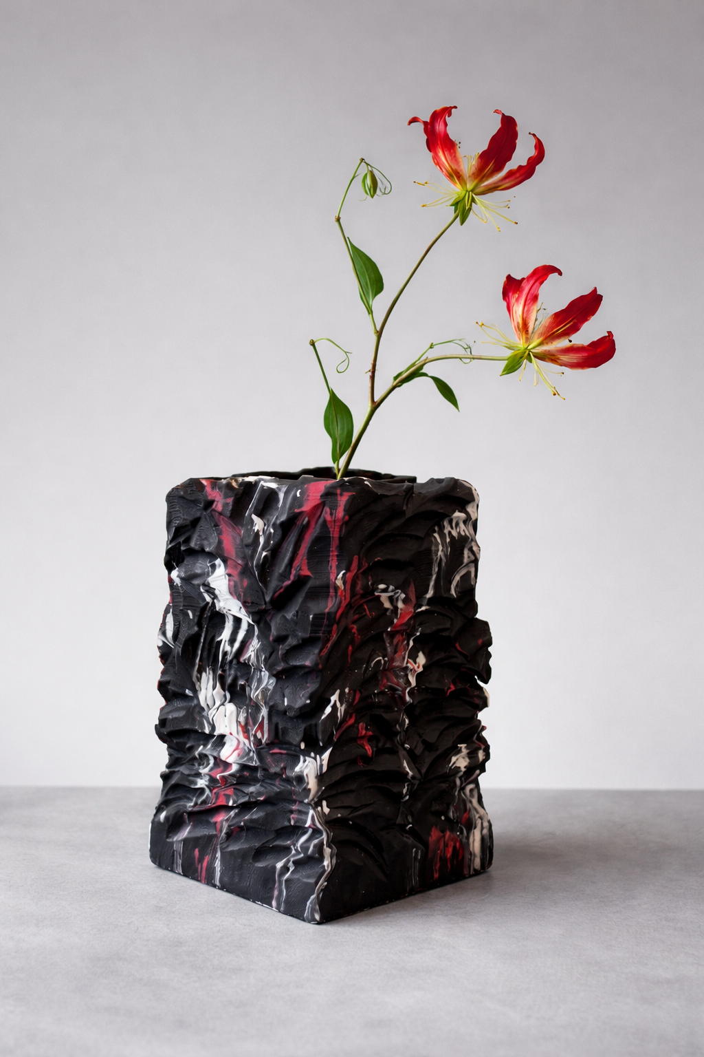 Een gestructureerde, zwarte, rechthoekige vaas met rode en witte verfspatten, gevuld met twee exotische rode en gele bloemen.