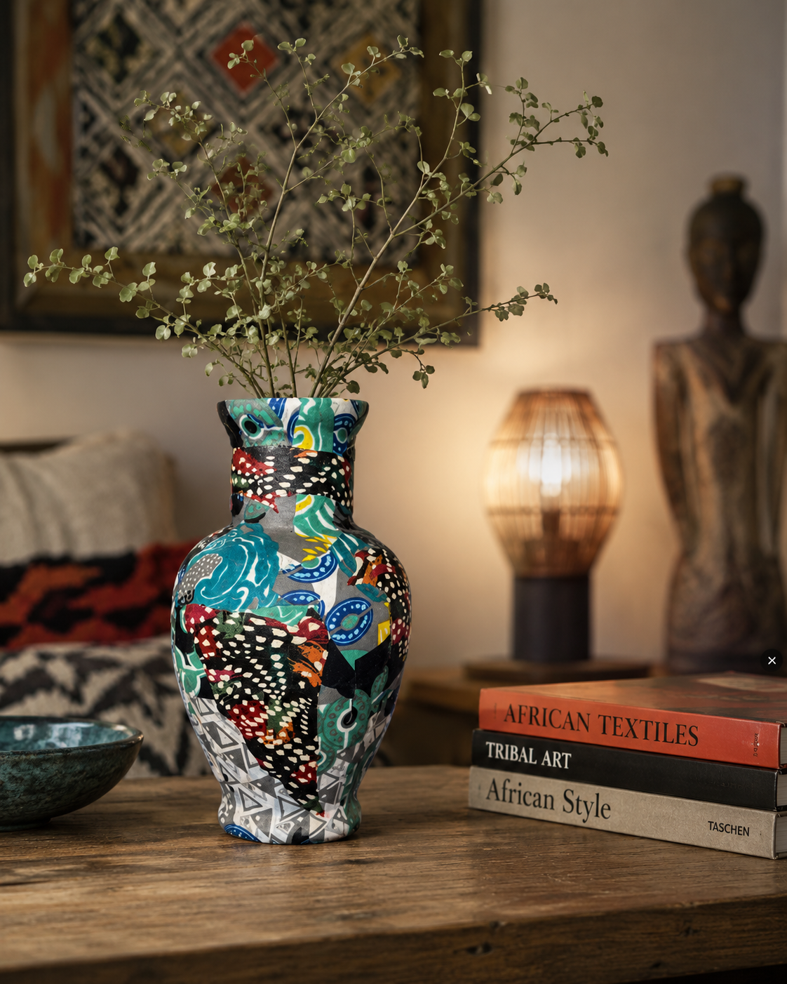 Een kleurrijke mozaïekvaas met takken staat op een houten tafel in een gezellige woonkamer, naast opgestapelde boeken en decoratie.