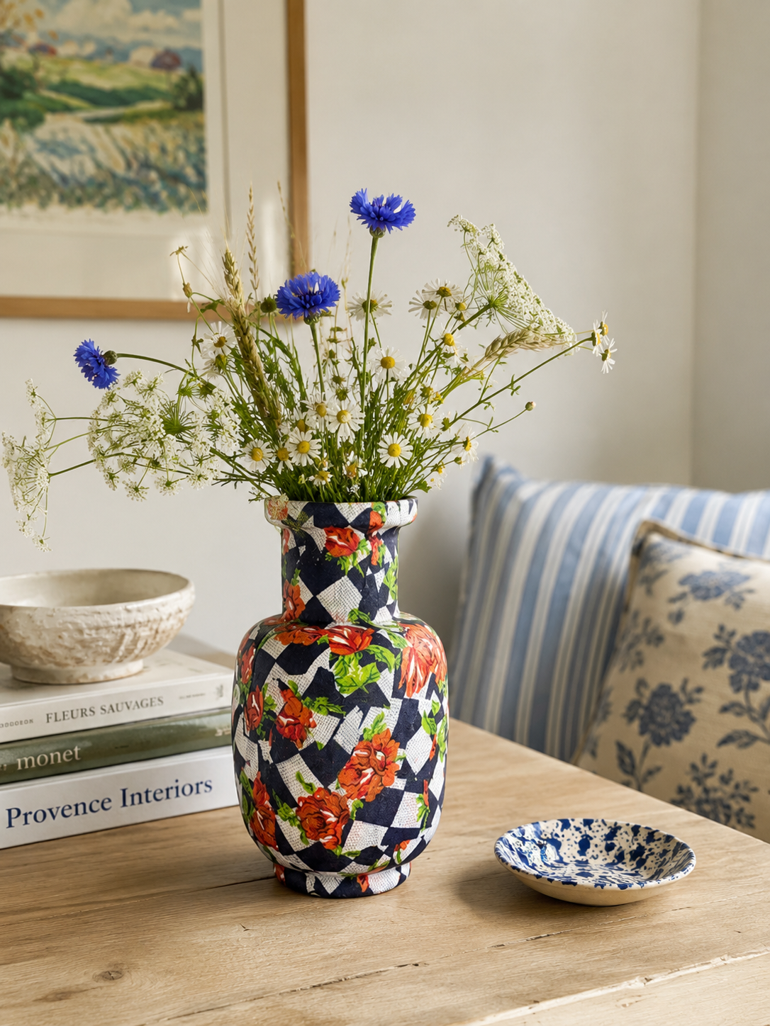 Mozaïekvaas met blauwe bloemen op een houten tafel naast een gestreepte bank en kleine schaaltjes.