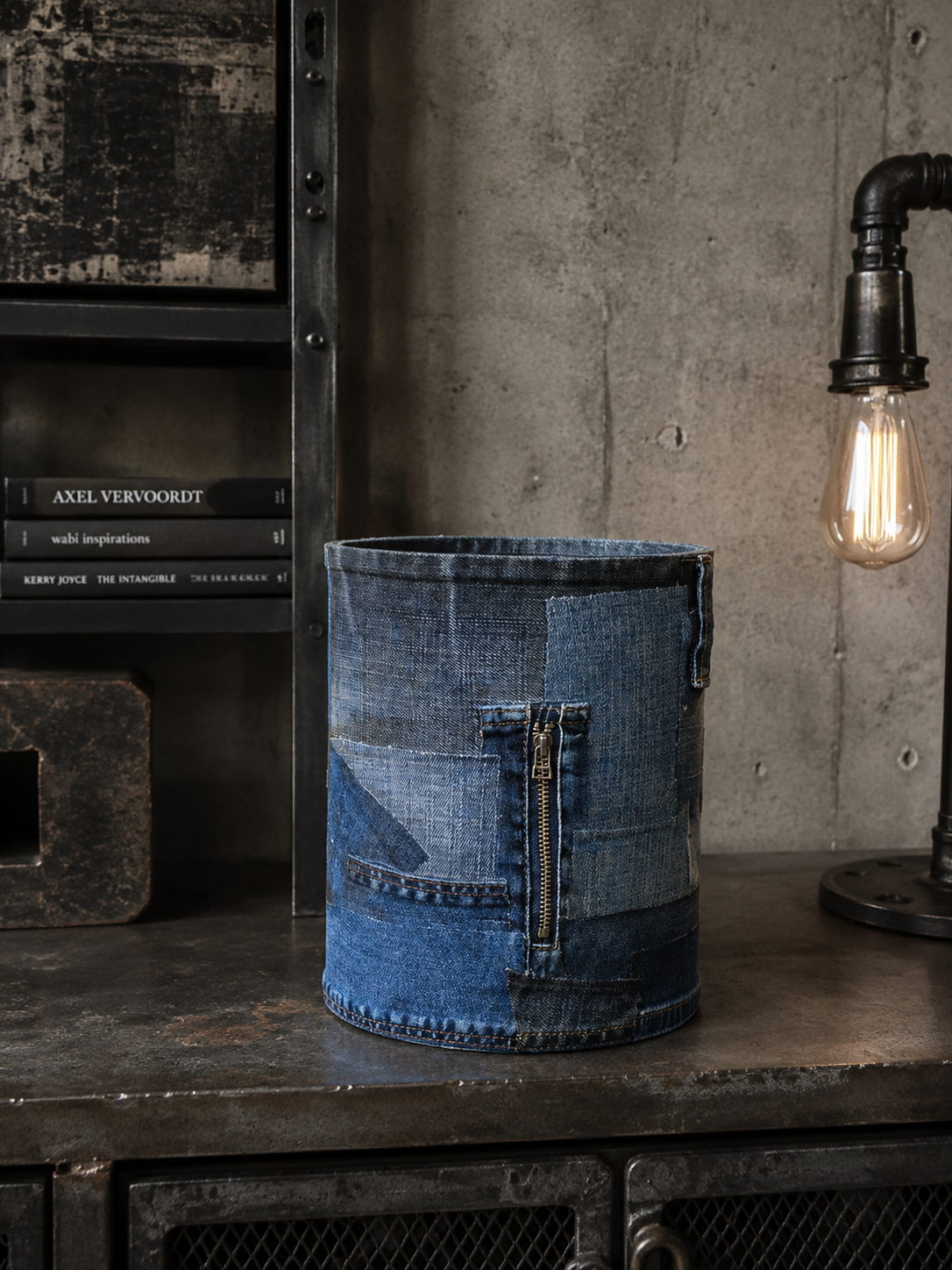 Een emmer van blauwe denim staat op een donkere plank naast een gloeiende Edison-lamp en een betonnen muur.