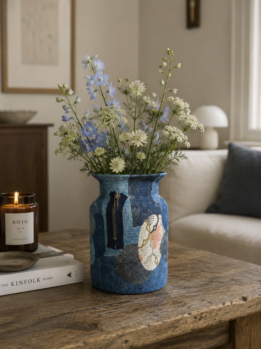 Een blauwe keramische vaas met wilde bloemen staat op een rustieke salontafel naast een brandende kaars en boeken.