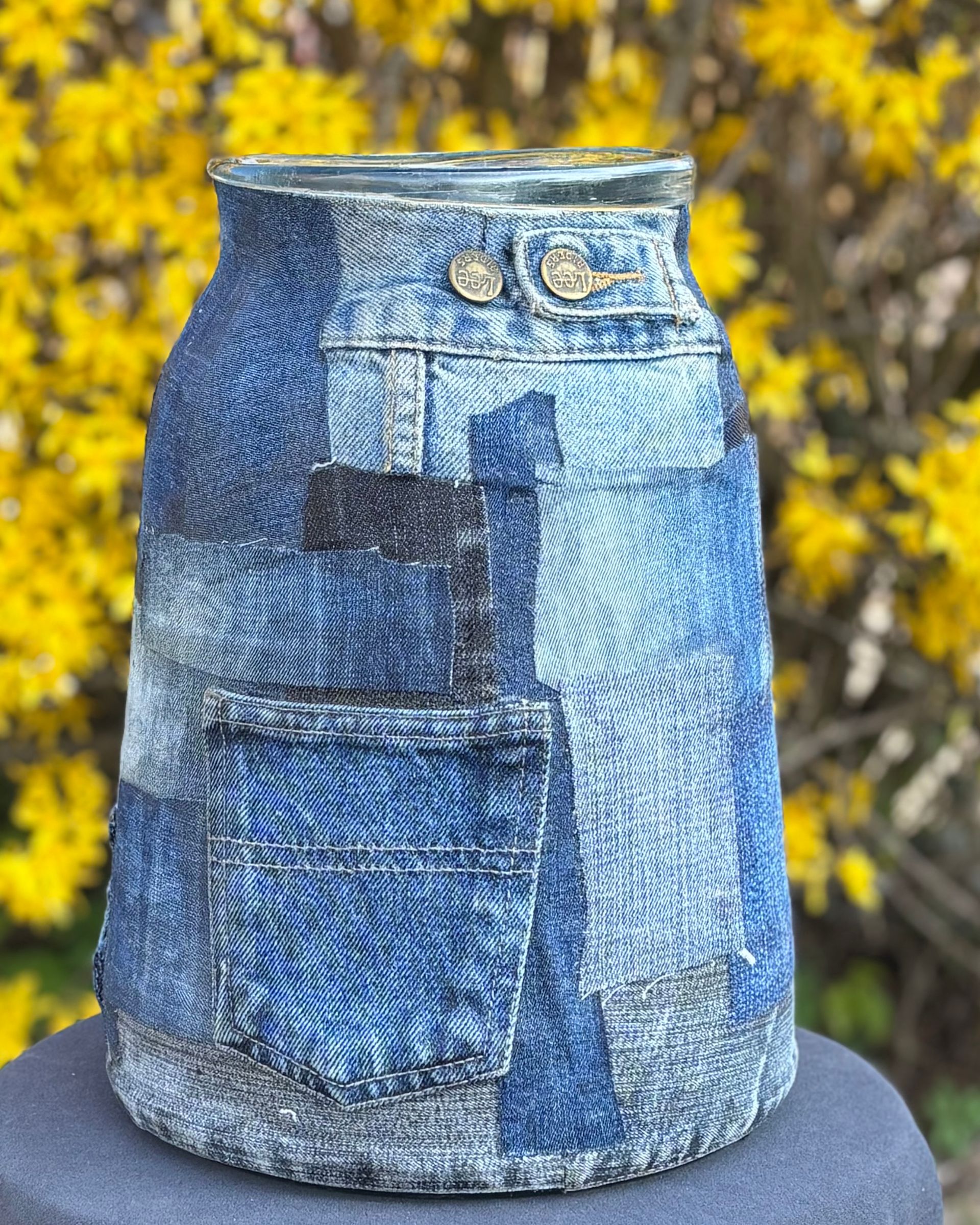 Een glazen vaas, hergebruikt met een patchwork hoes van blauwe denimstof, voorzien van een zakje en metalen knopen, tegen een achtergrond van gele bloemen.