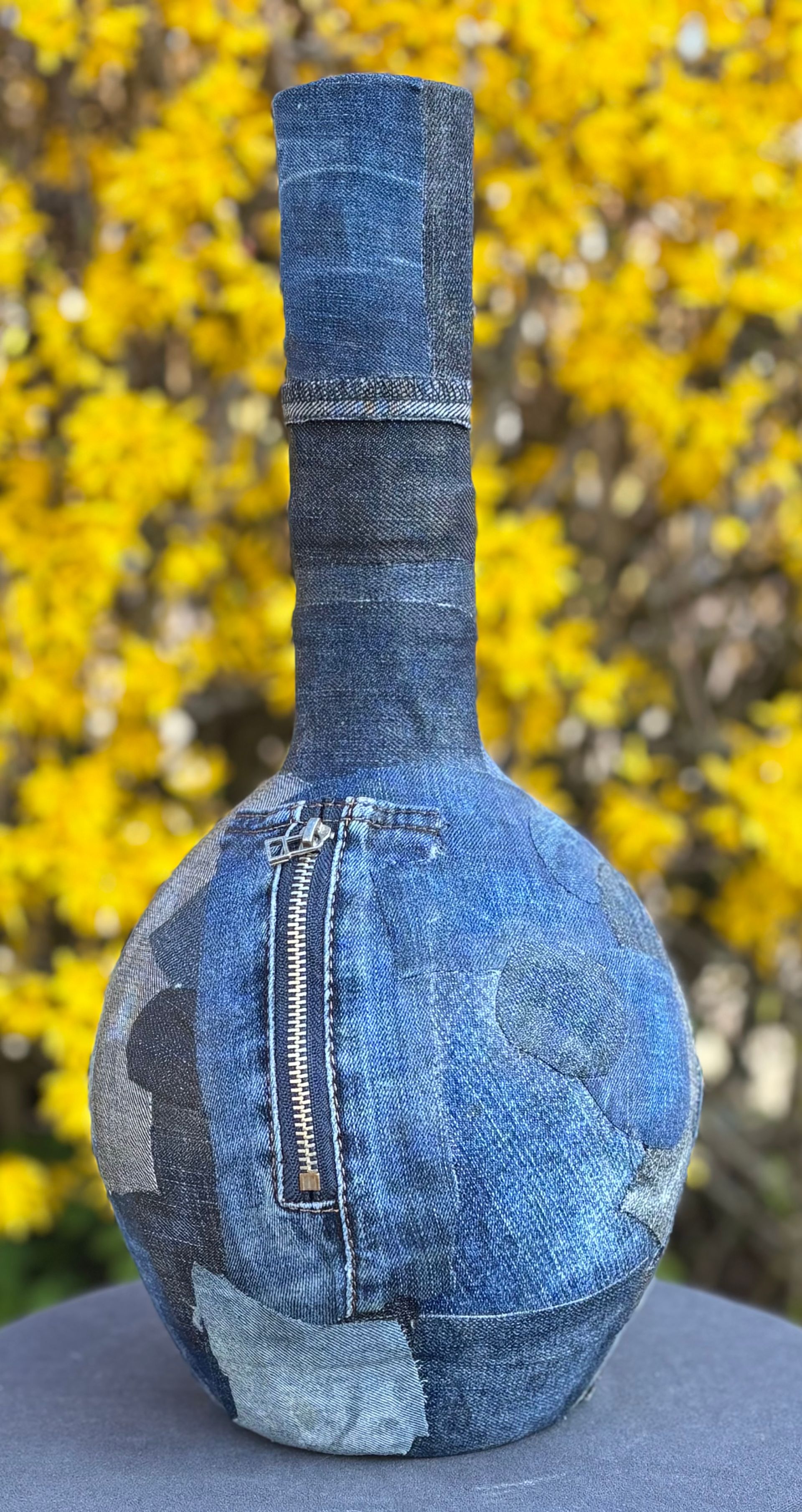 Een vaas met een structuur, bekleed met blauwe denimstof en voorzien van een opvallende verticale rits, geplaatst tegen een achtergrond van gele bloemen.