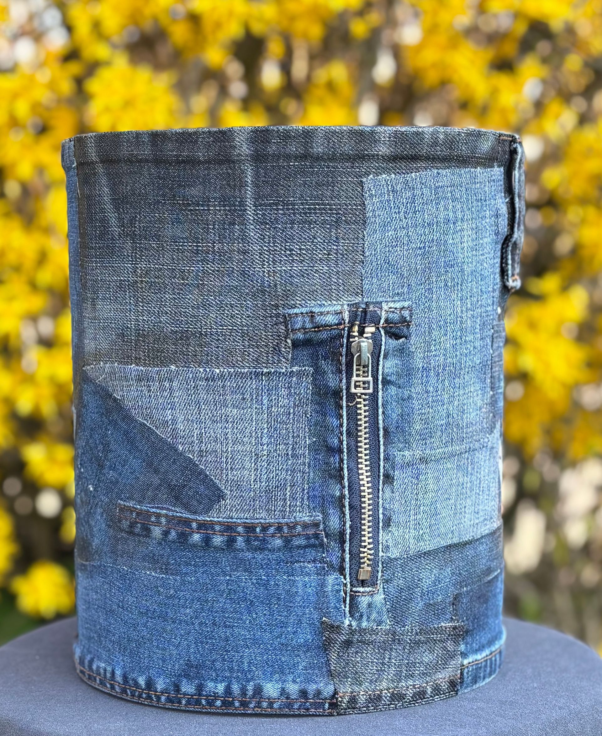 Een cilindervormige bloempot van denim patchwork met een functionele metalen rits, geplaatst tegen een wazige achtergrond van gele bloemen.
