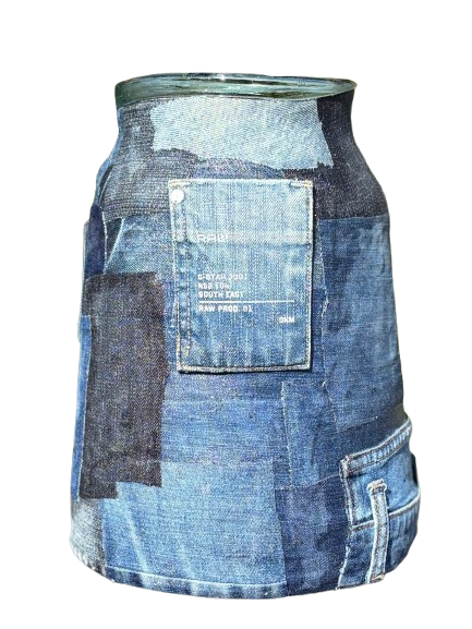 Een glazen vaas, bedekt met blauwe denim patchworkresten, met een zakje en metalen knoopjes als accenten, tegen een achtergrond van gele bloemen.