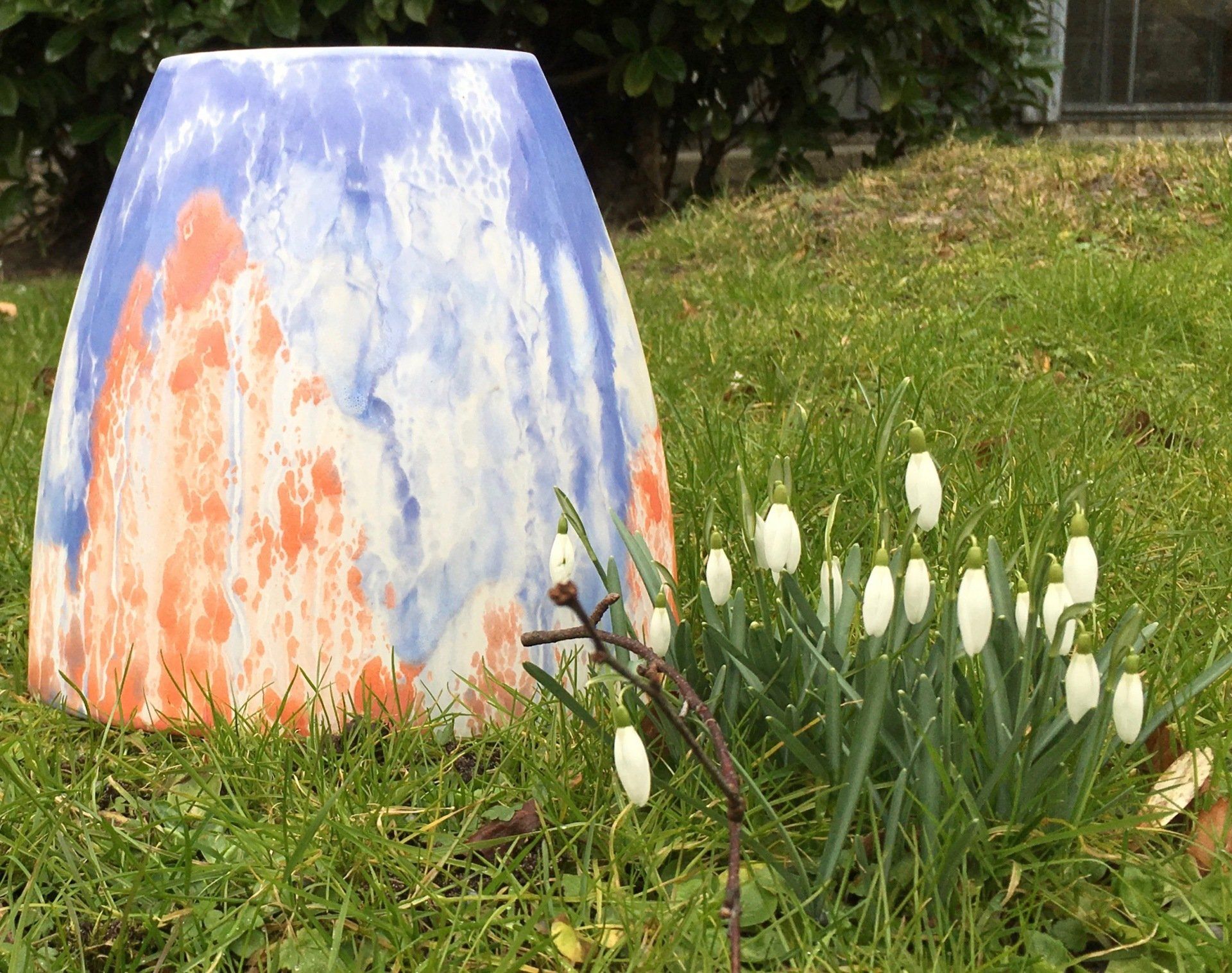 Een blauw en oranje vaas staat in het gras naast wat witte bloemen.