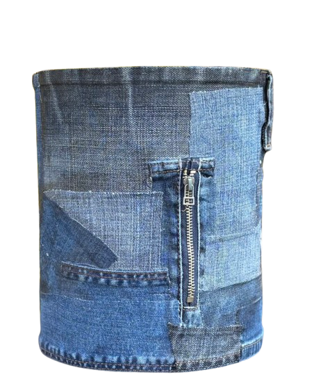 Een cilindervormige plantenbak van blauwe patchworkdenim met een verticale metalen ritssluiting, geplaatst tegen een wazige gele struik.