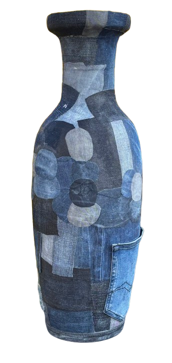 Een vaas van patchwork blauwe denimstof staat op een grijze sokkel tegen een achtergrond van gele bloeiende struiken.