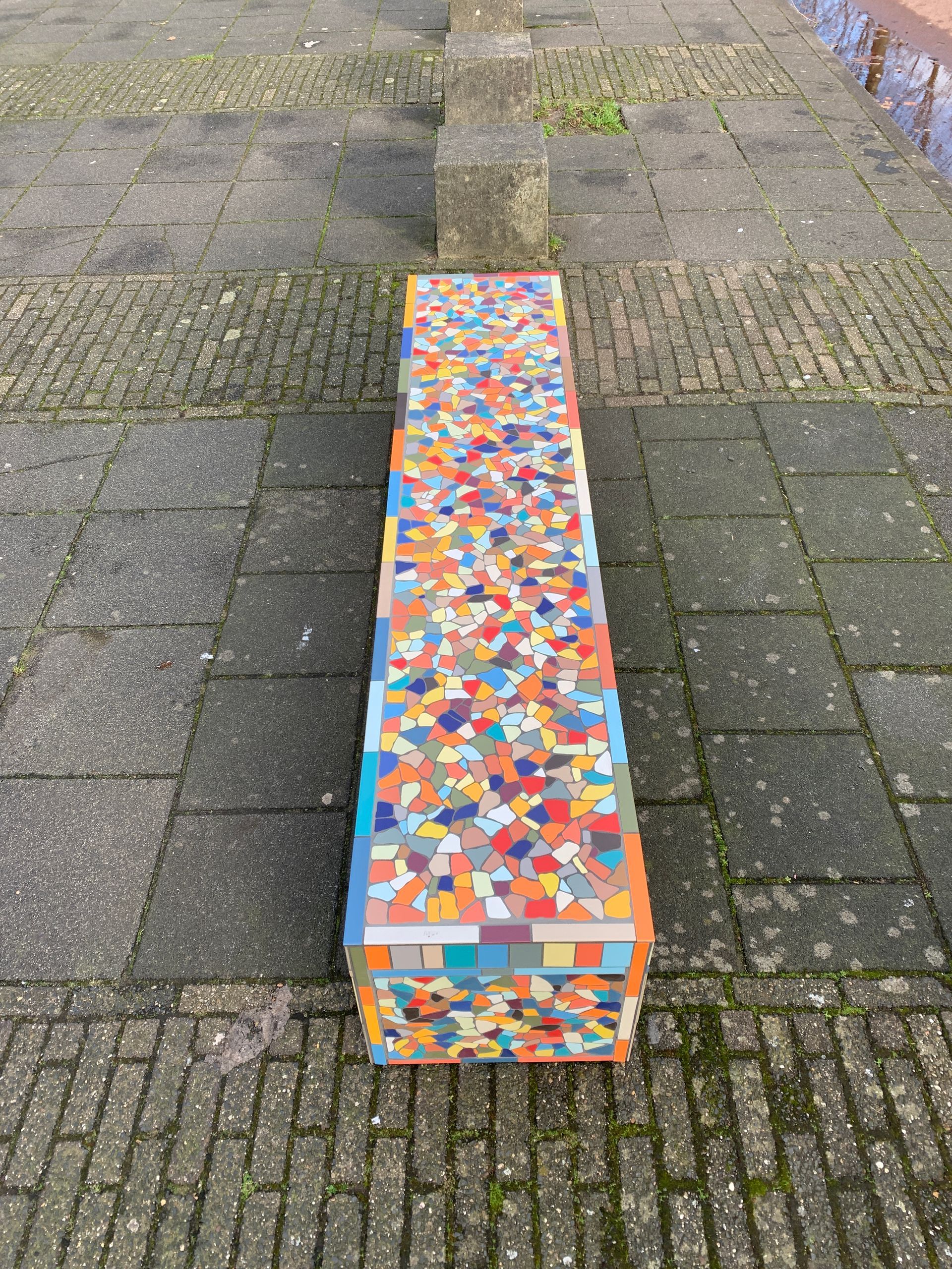 Een lange kleurrijke bank staat op een stoep van bakstenen.