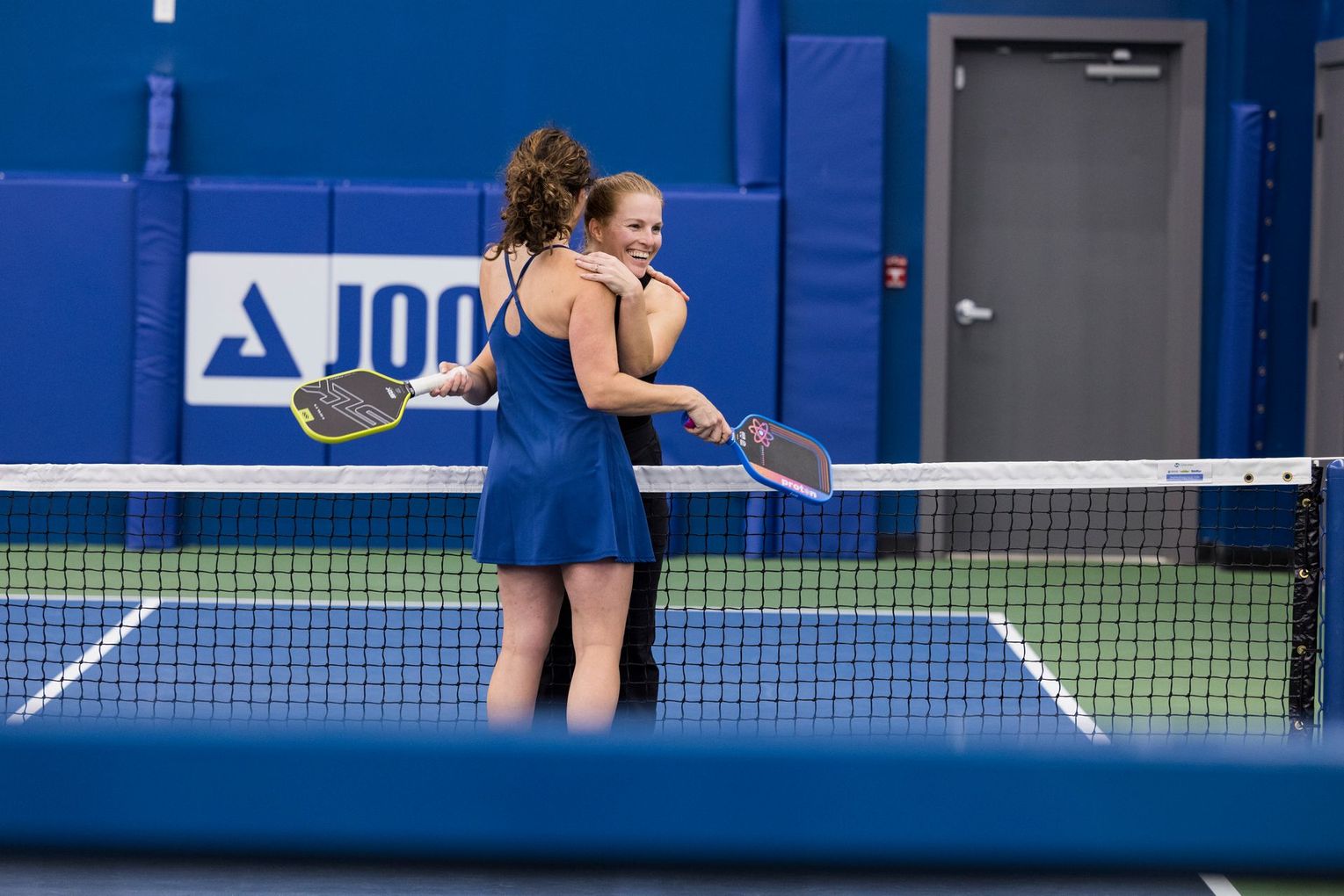 Two pickleball players in blue athletic wear hug over the net on an indoor court with a blue wall and logo.