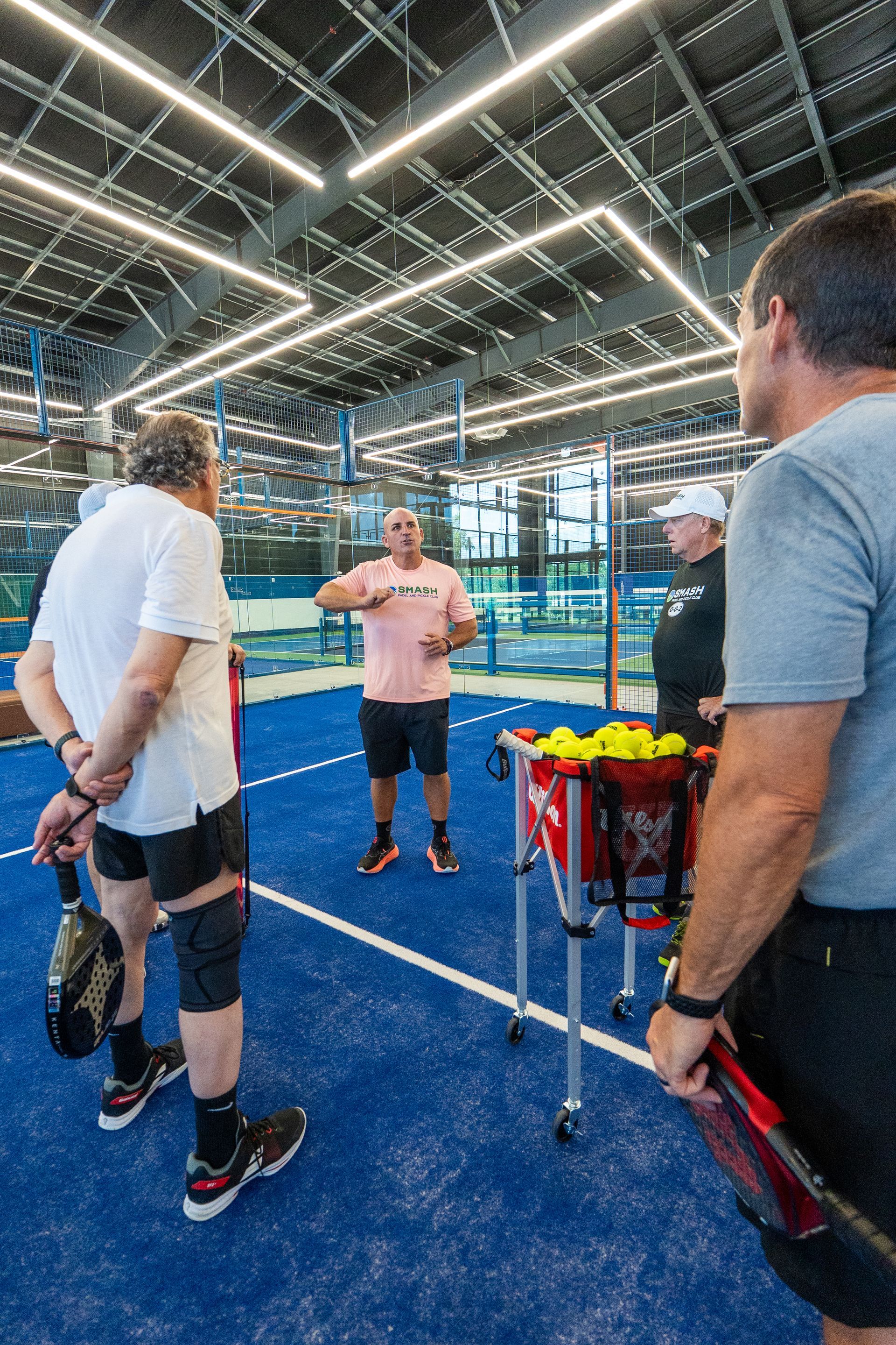 Group of people on a blue padel court, listening to a man in a pink shirt, with a ball cart nearby.