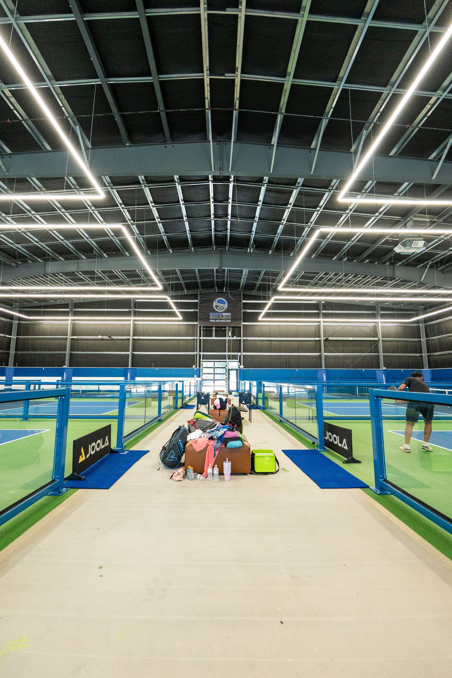 Indoor sports facility; people near tennis/pickleball courts. Overhead lighting, blue courts, and gear.