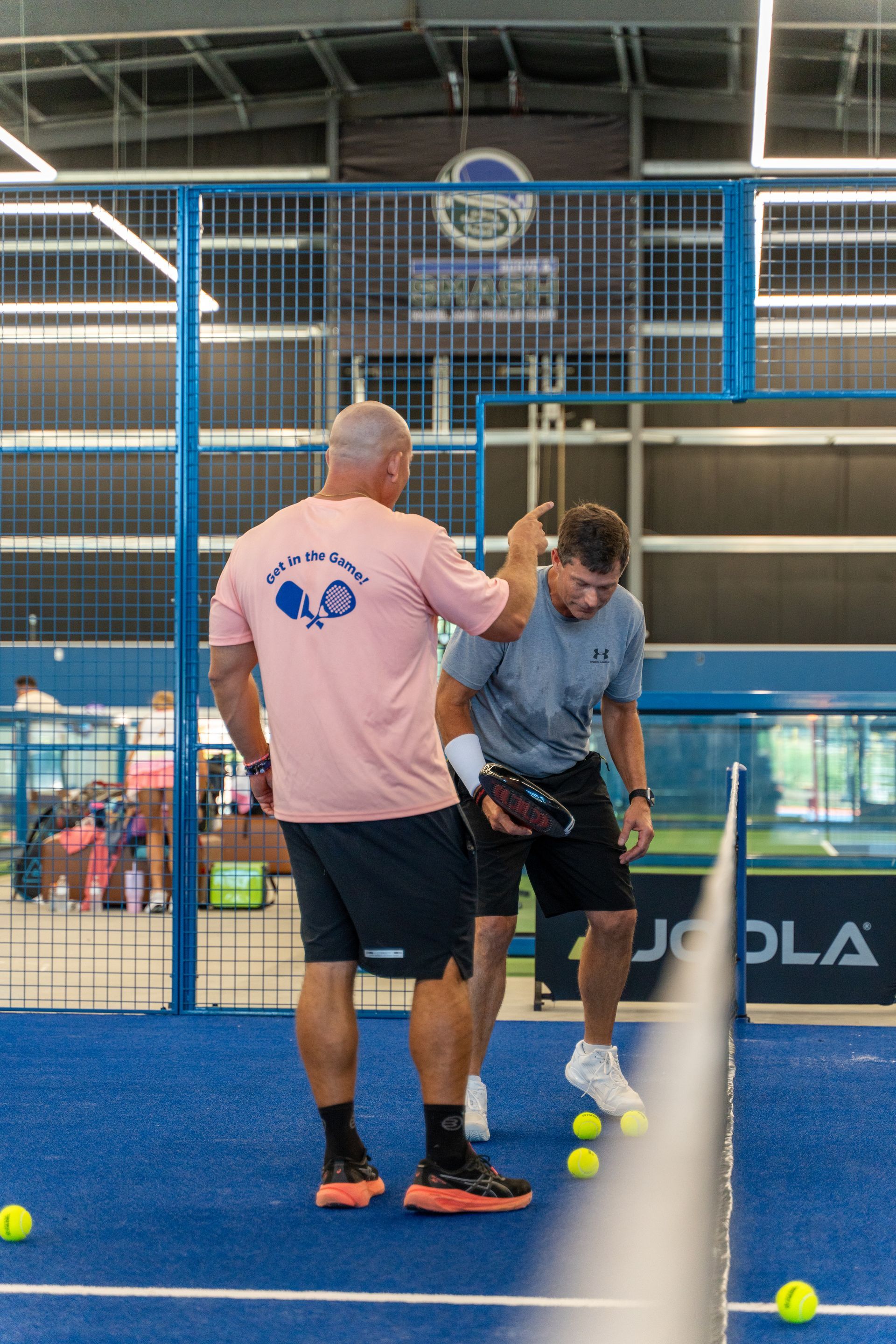 Two men on a blue padel court. One man in pink shirt instructs a player holding a racket, with yellow balls nearby.