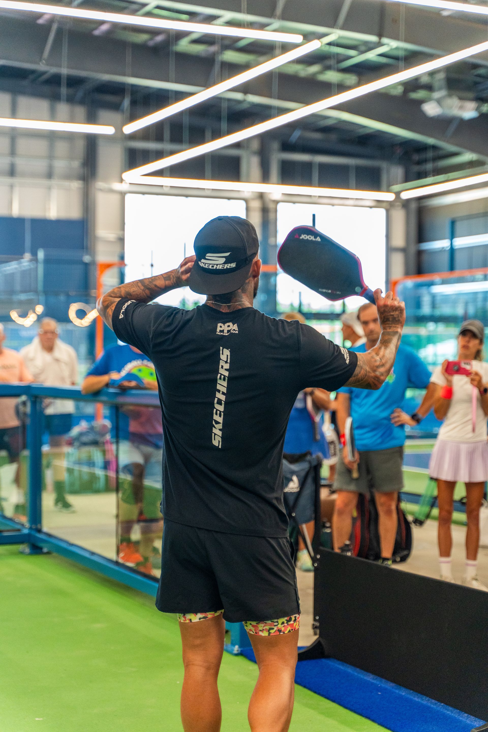 Man in black gear with a paddle raising his arm, on a court, other players visible.