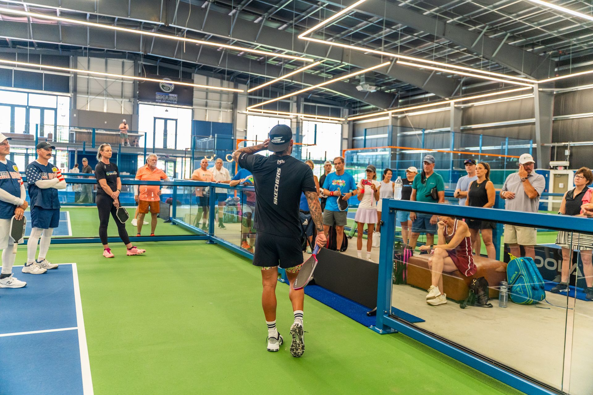 Indoor pickleball court with players. Man in black shirt gestures to a crowd. Green court, blue walls.