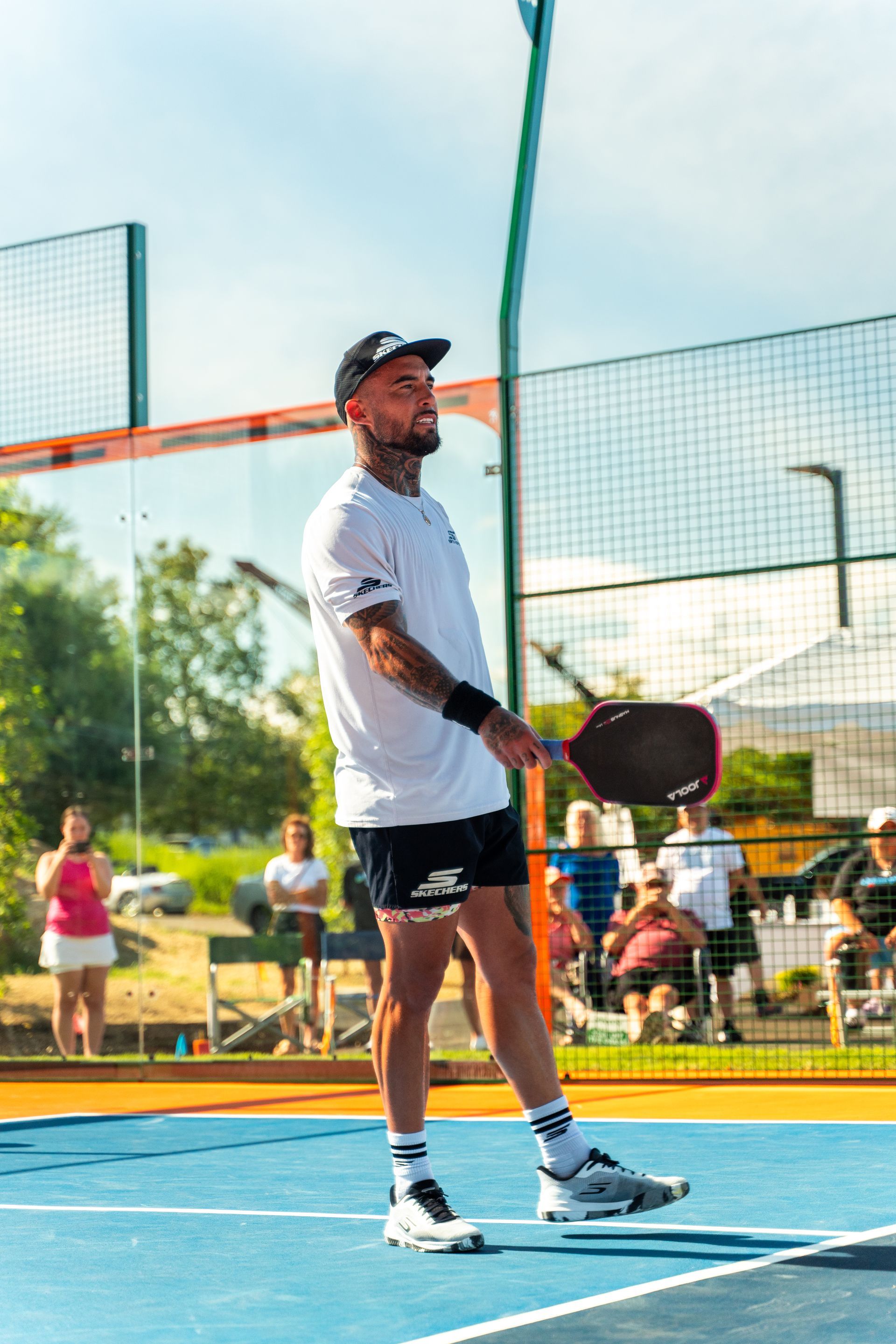 Man with tattoos playing pickleball on a blue court, looking up, holding a paddle.