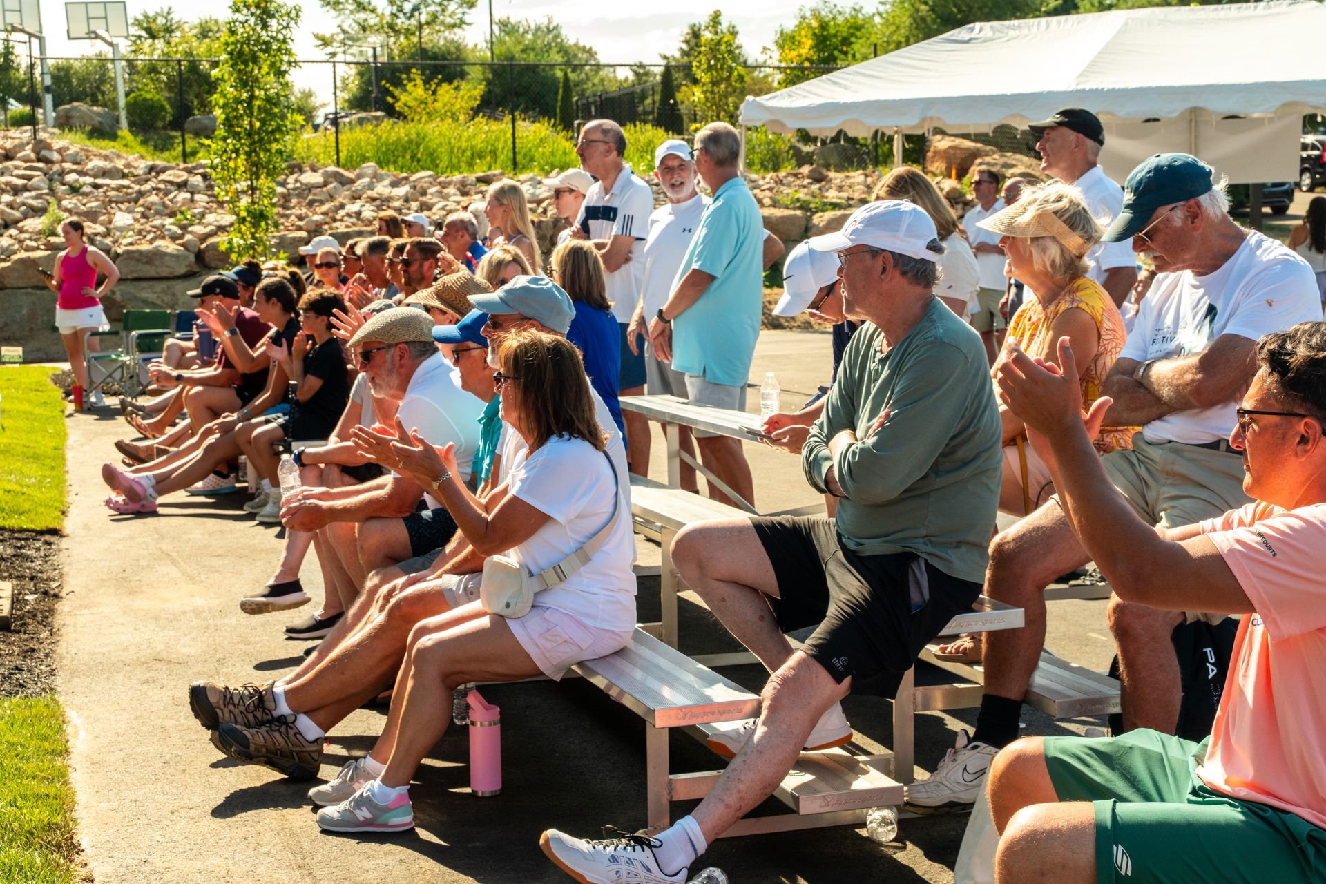 Spectators seated on benches, clapping and watching an outdoor event on a sunny day.