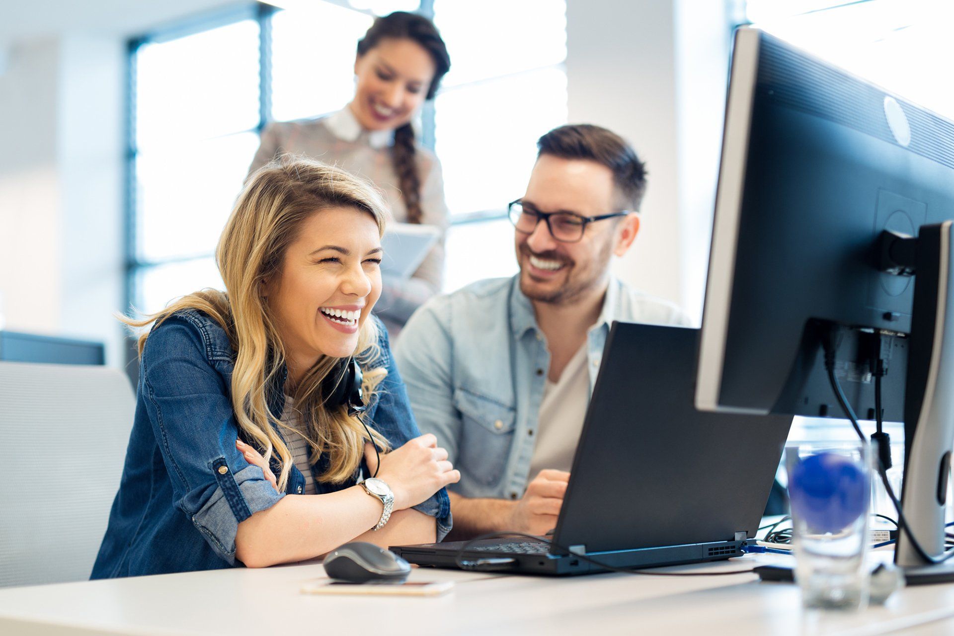 Three smiling office workers collaborating on a laptop and computer in a modern office setting.