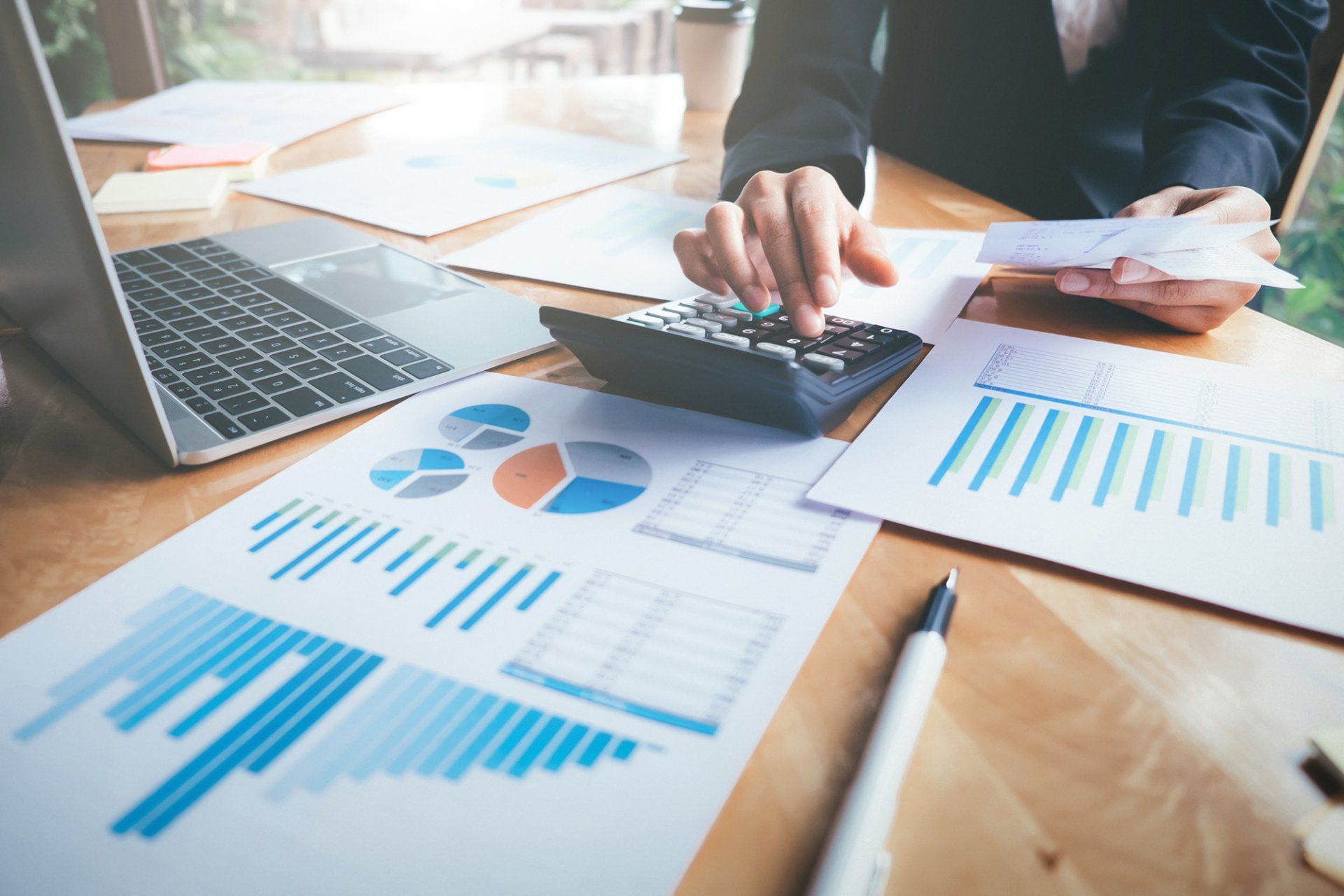 Person in a business suit using a calculator, reviewing financial reports with charts and a laptop on a wooden desk.