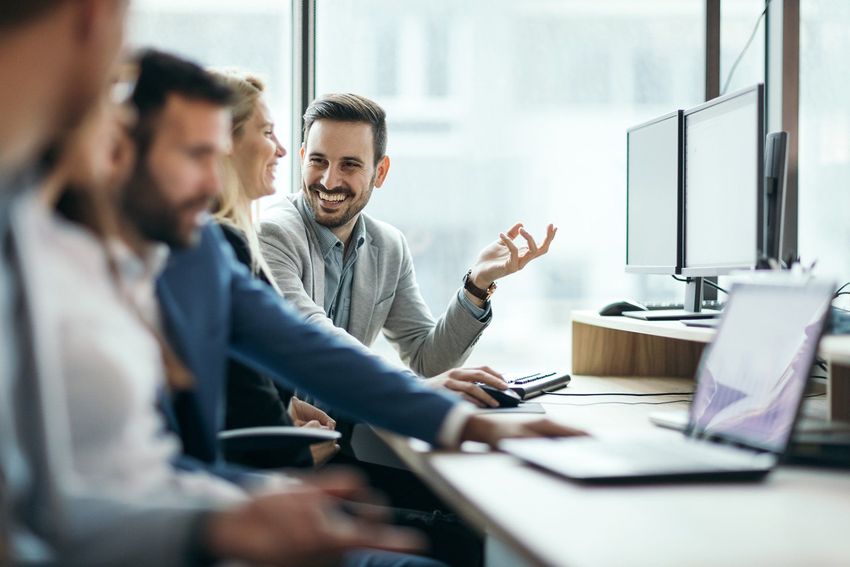 Four people collaborating at a desk in an office. Man smiles, gestures with his hand. Windows in background.