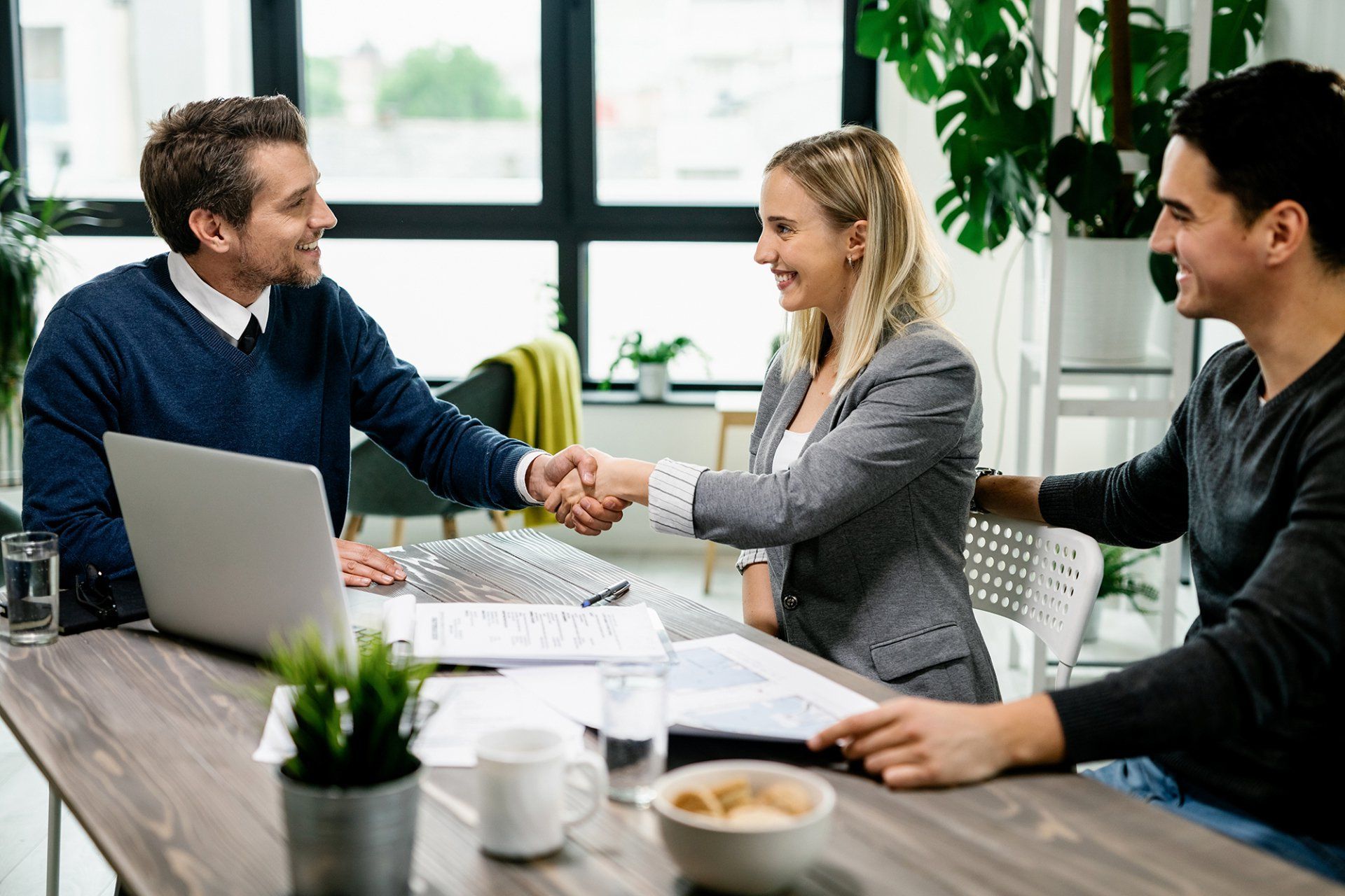 Three people in an office shake hands over a desk, smiling. A laptop and plants are visible.