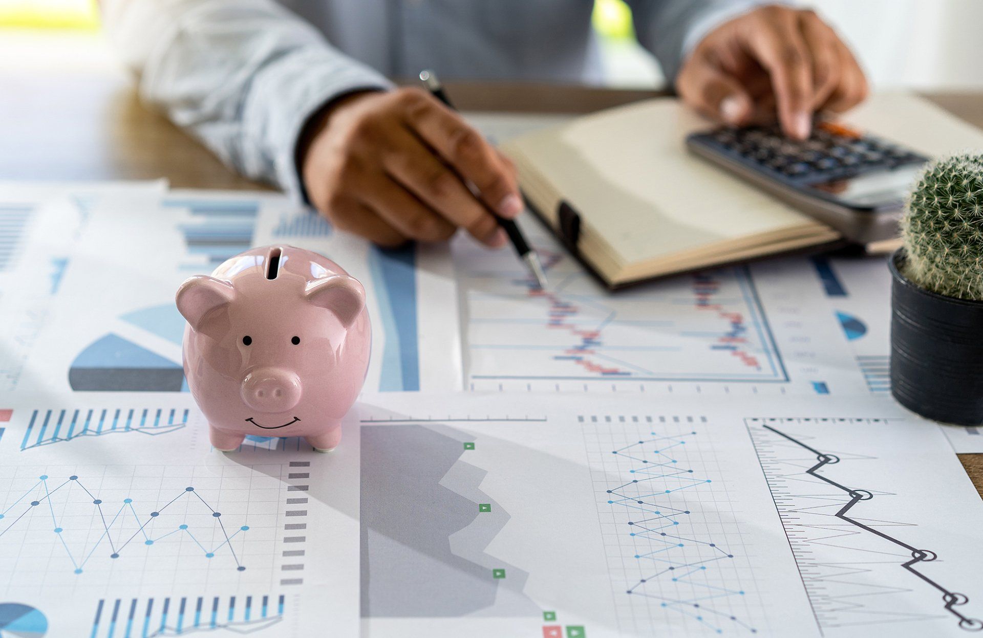 Person analyzing financial charts with a piggy bank, calculator, and notepad on a desk.