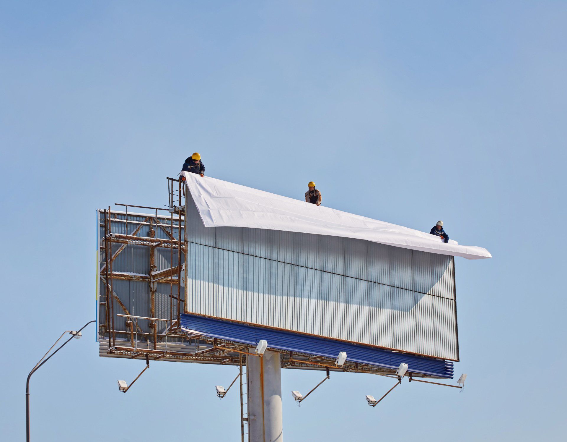 Workers in yellow hard hats installing white billboard sheet on metal structure.