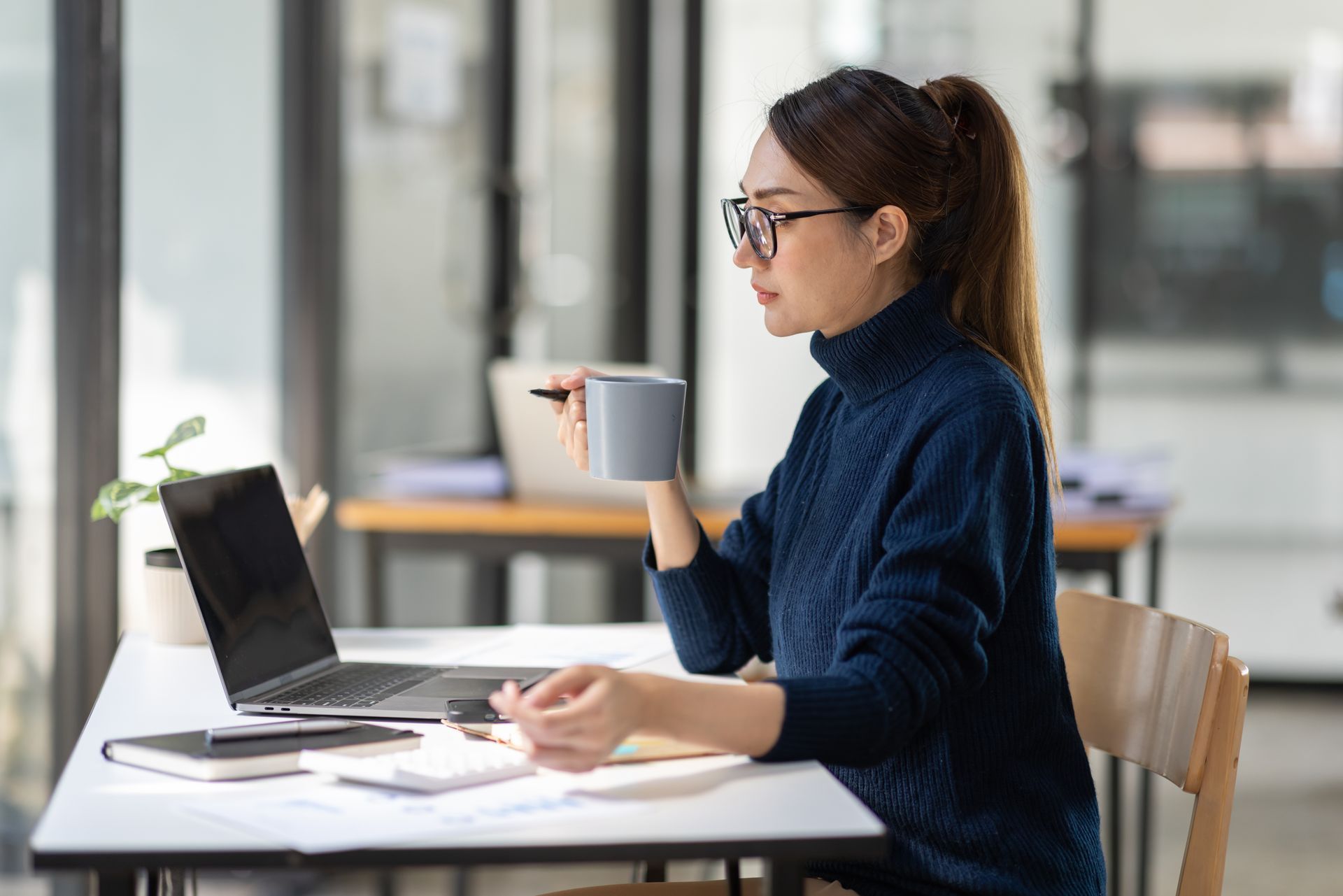 Woman with glasses working on laptop, sipping from mug at a desk, in office setting.
