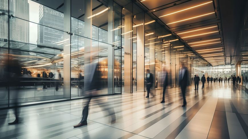 Blurred figures in suits walk through a modern glass-walled building with bright lighting.