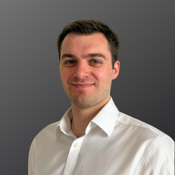 Man with a light beard, smiling, wearing a blue striped button-down shirt, grey background.