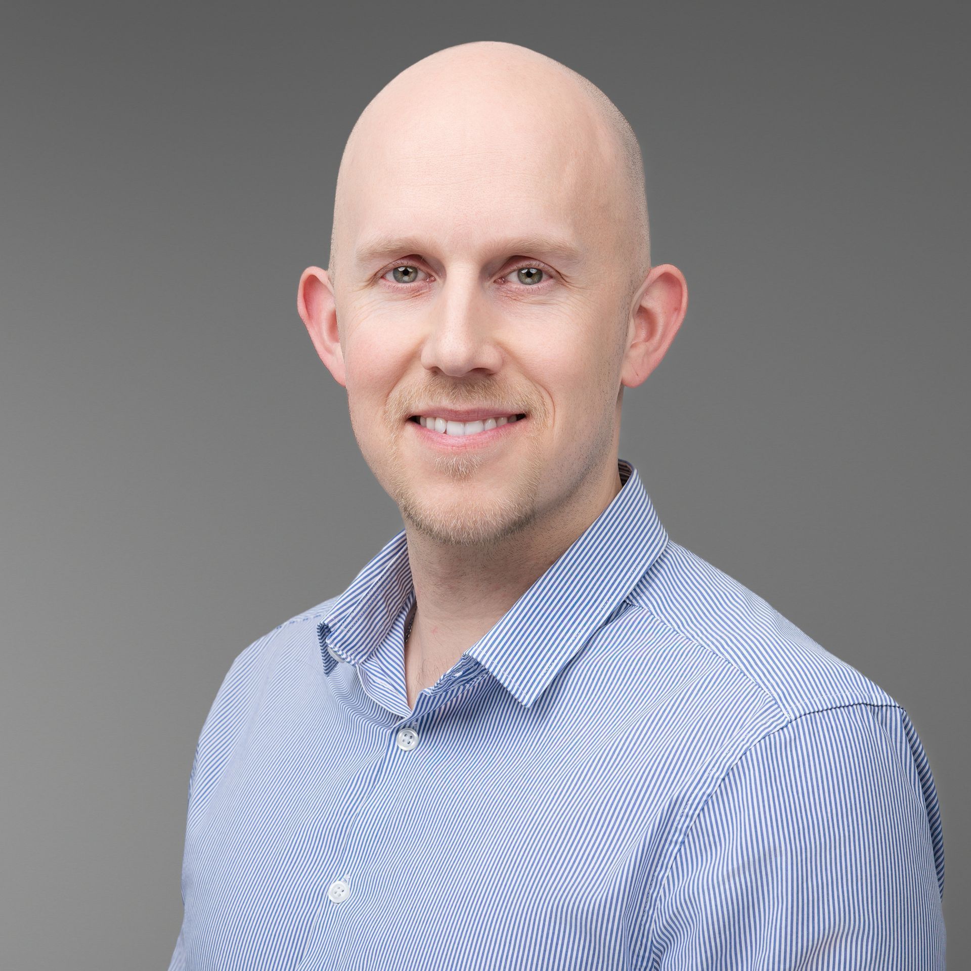 Man with a light beard, smiling, wearing a blue striped button-down shirt, grey background.