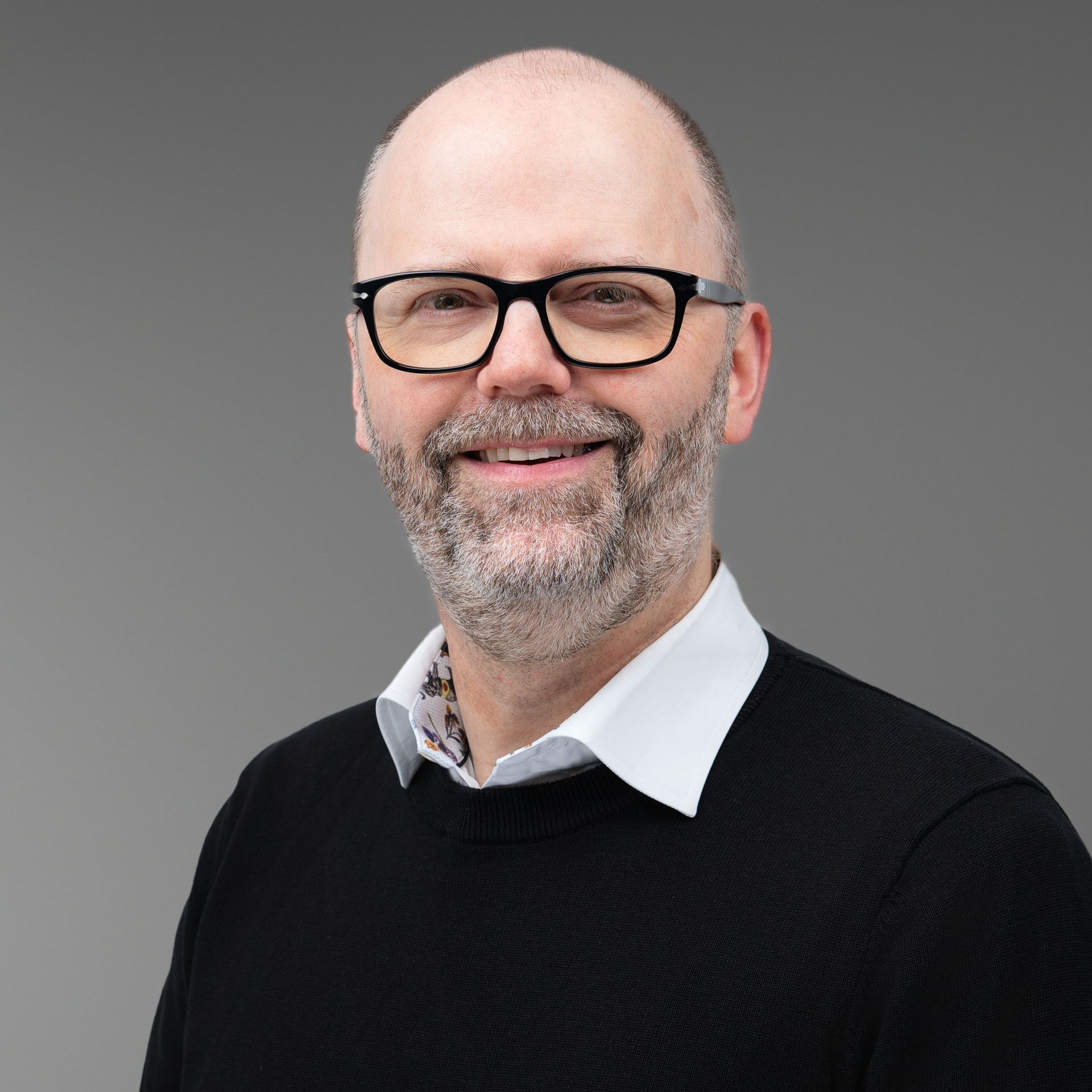 Man with glasses and beard, smiling, wearing black sweater over white collared shirt, gray background.