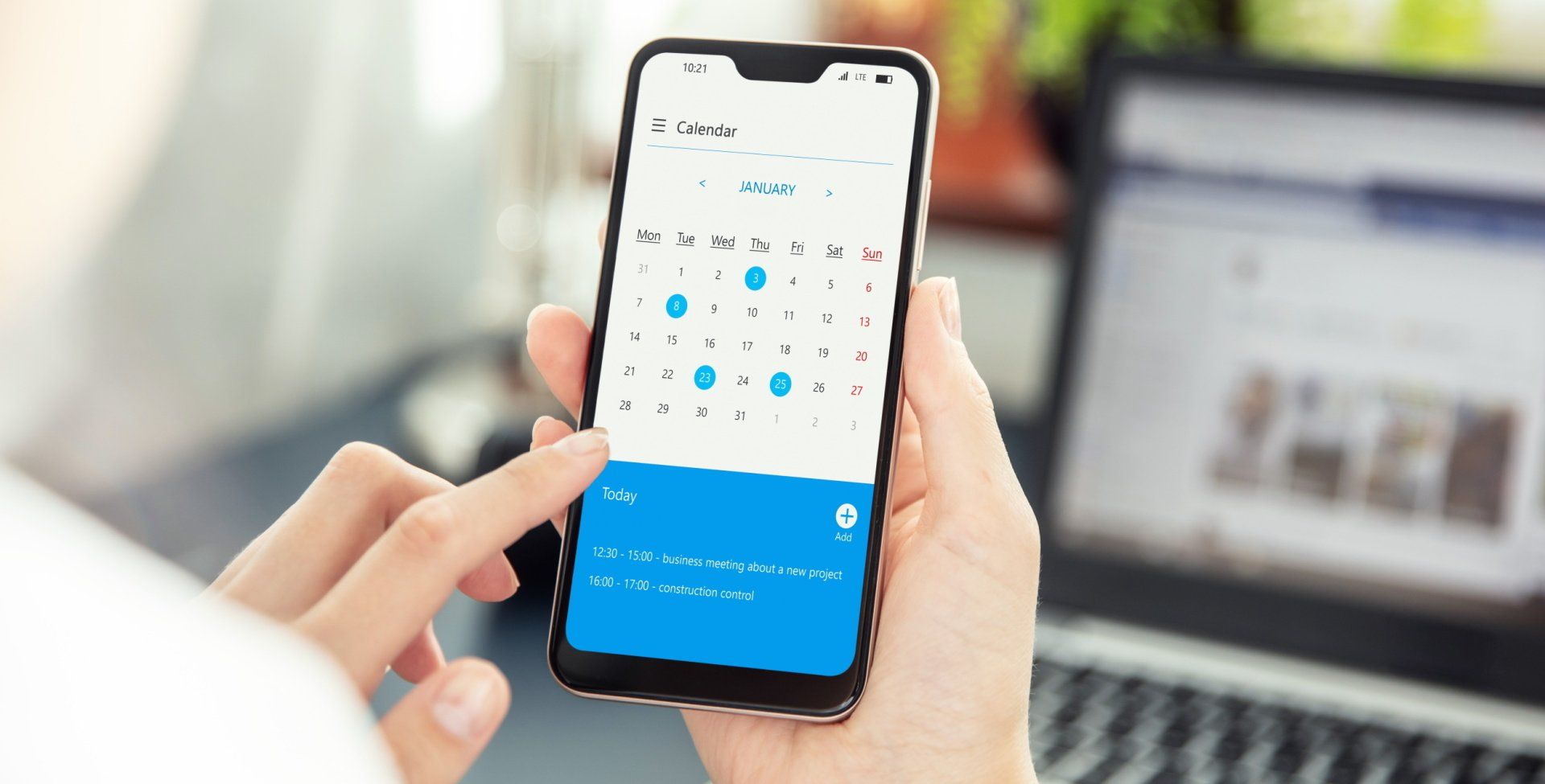 Person holding smartphone displaying a calendar, finger touching the screen, with a laptop in the background.