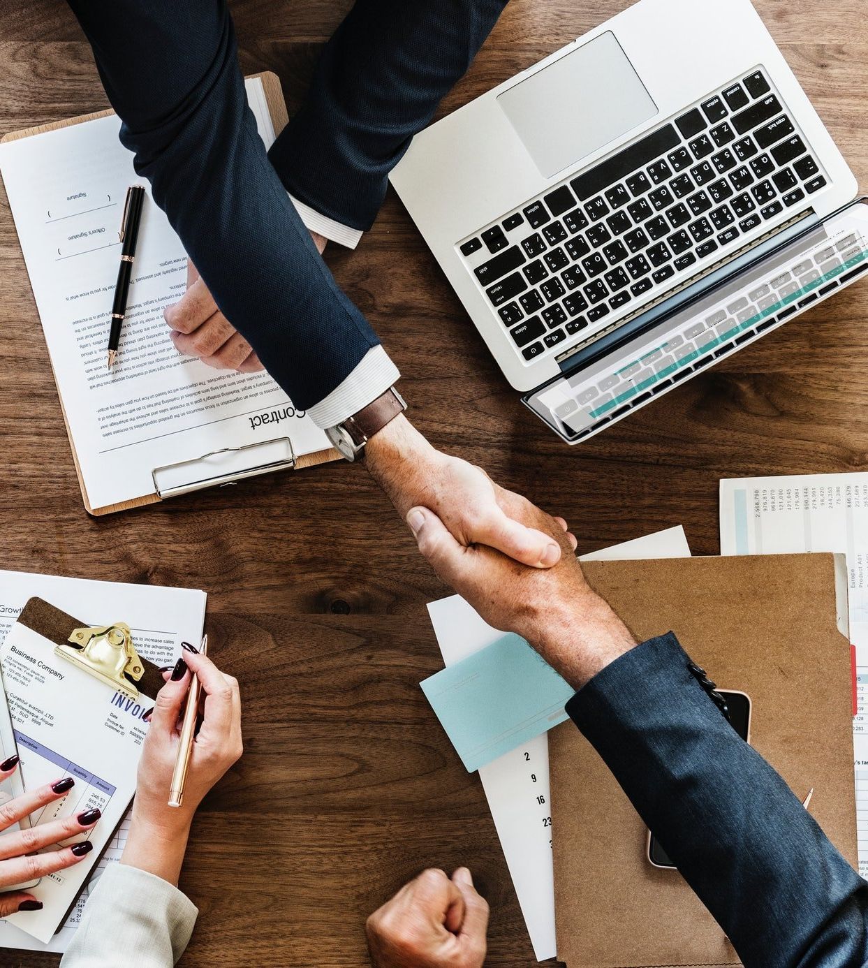 Two people in suits shaking hands over a table with papers and a laptop.