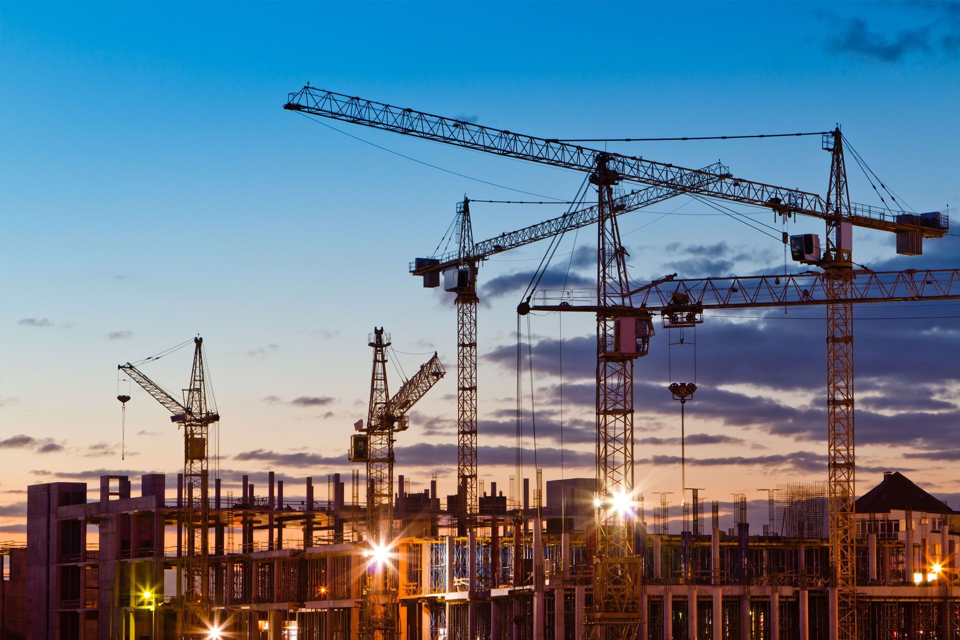Construction site at dusk with cranes silhouetted against a blue and orange sky, building frameworks lit up.