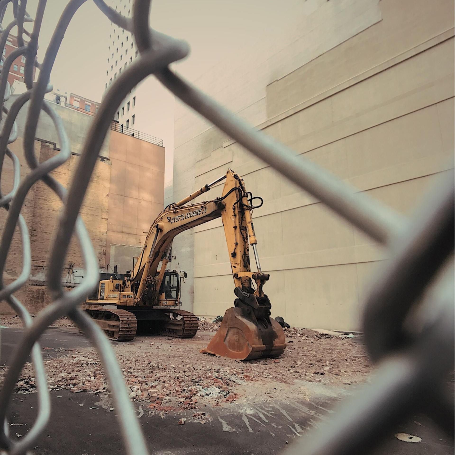 Yellow excavator behind a chain-link fence, at a construction site, against a beige building.