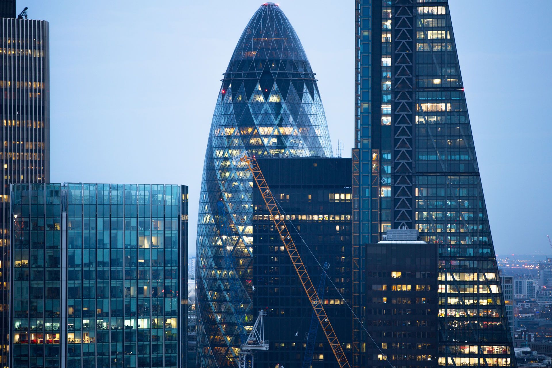 Skyscrapers in London's financial district at dusk, including the Gherkin, with city lights visible.