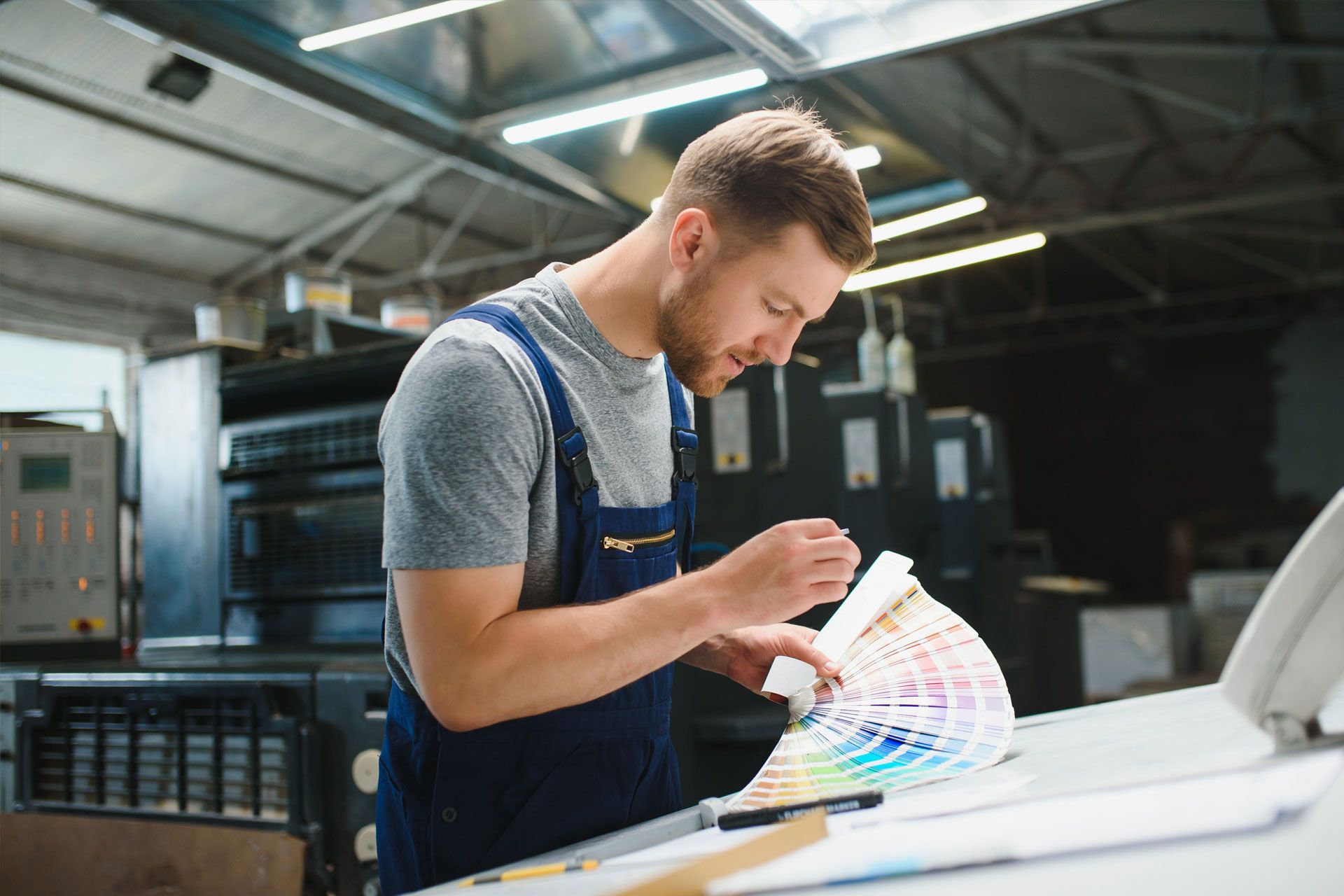 Man in blue overalls examines a color swatch fan in a printing shop.