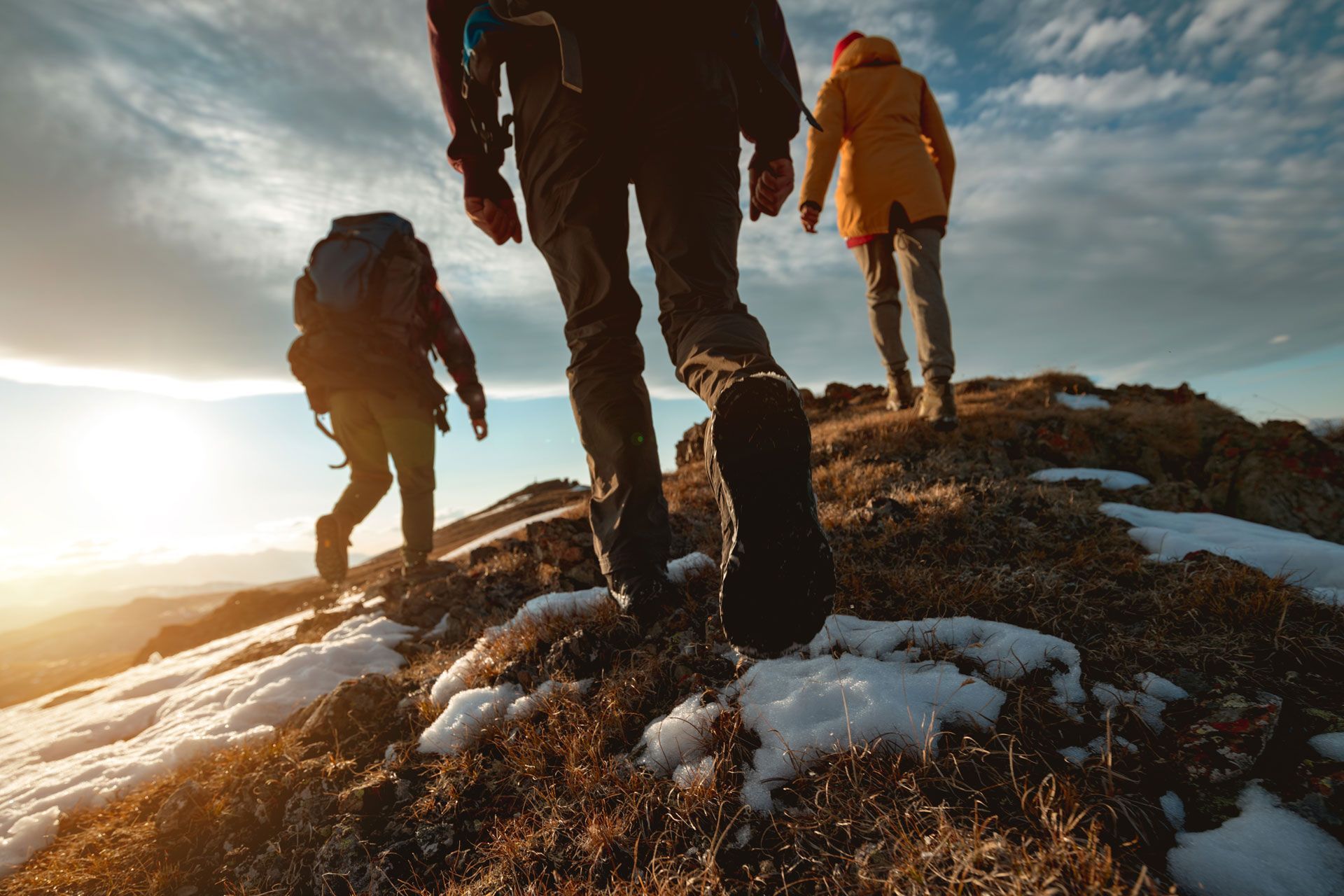 Hikers ascend a mountain path with patches of snow, backlit by a sunrise or sunset.