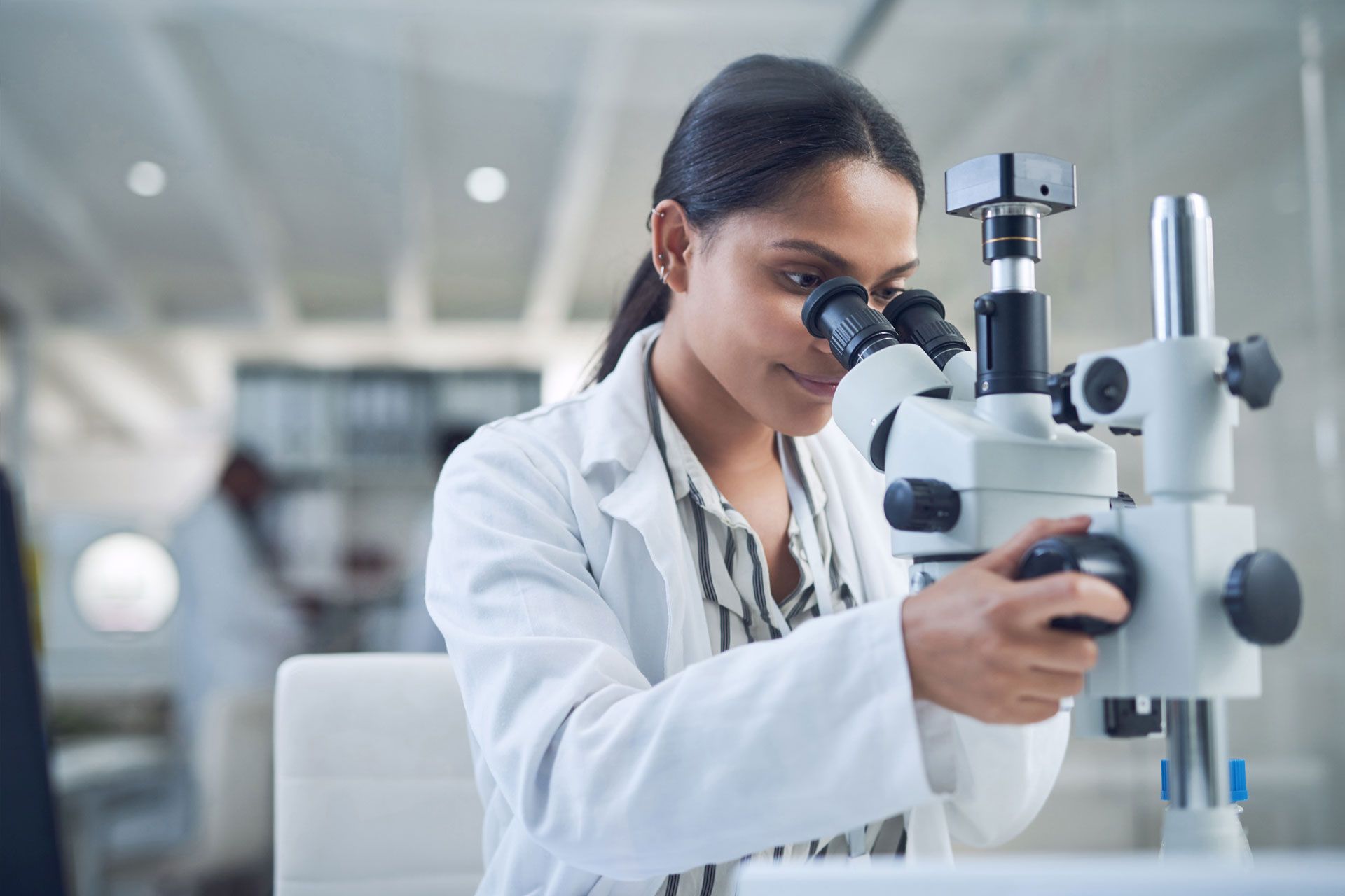 Woman in lab coat looking through microscope in a lab.