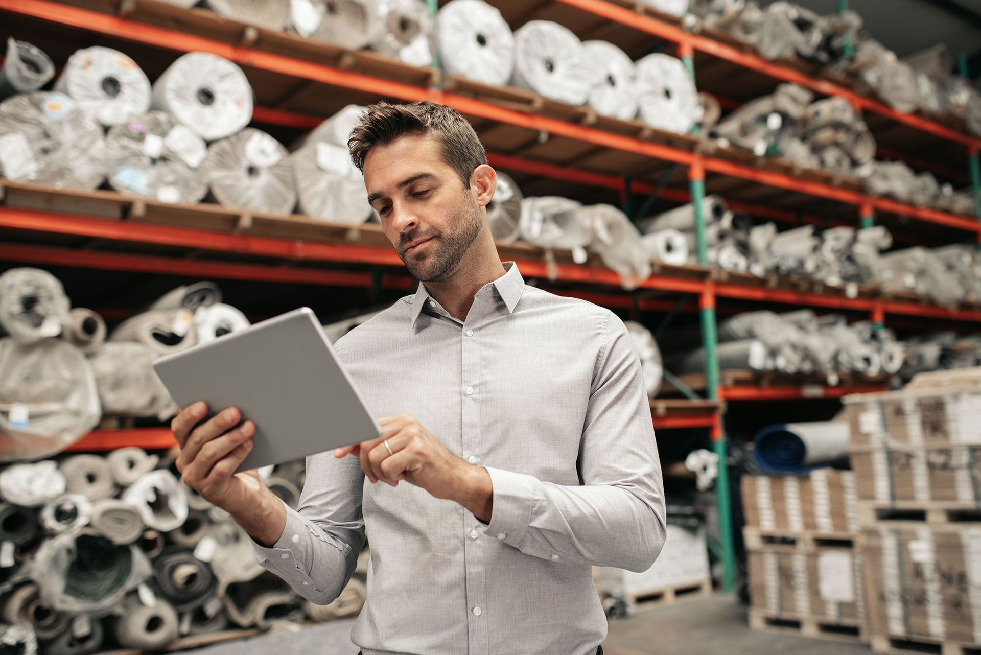 Man using a tablet in a warehouse, checking inventory; shelves filled with rolled items.