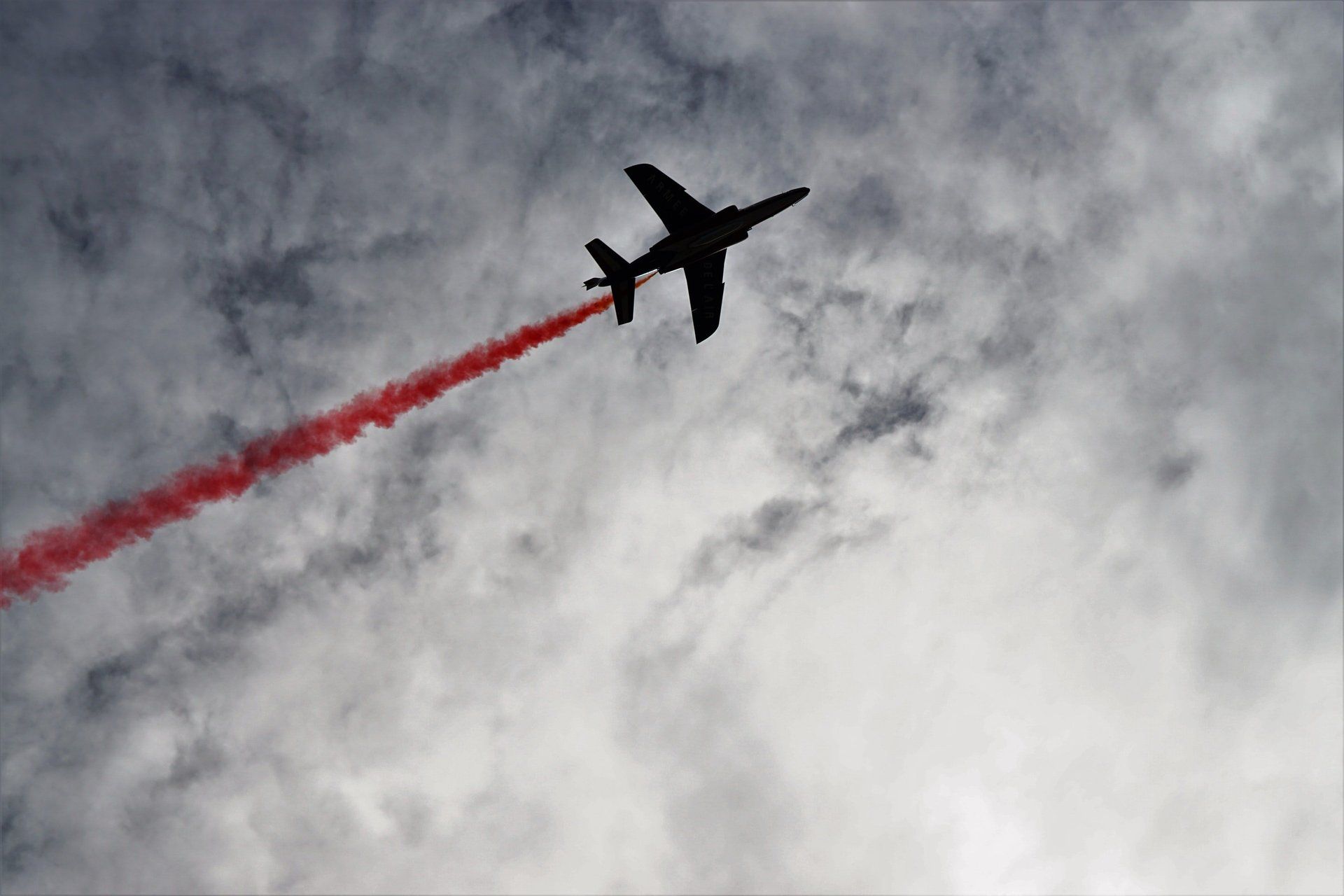 Silhouette of a jet plane streaking red smoke against a cloudy sky.