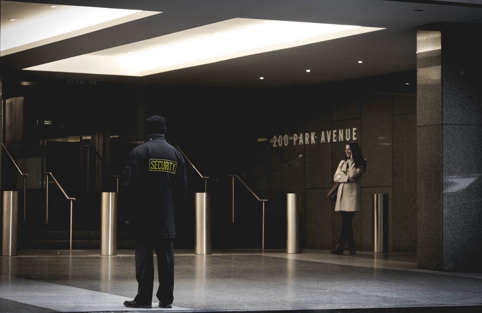 Security guard stands near the entrance of 200 Park Avenue as a woman in a trench coat waits.