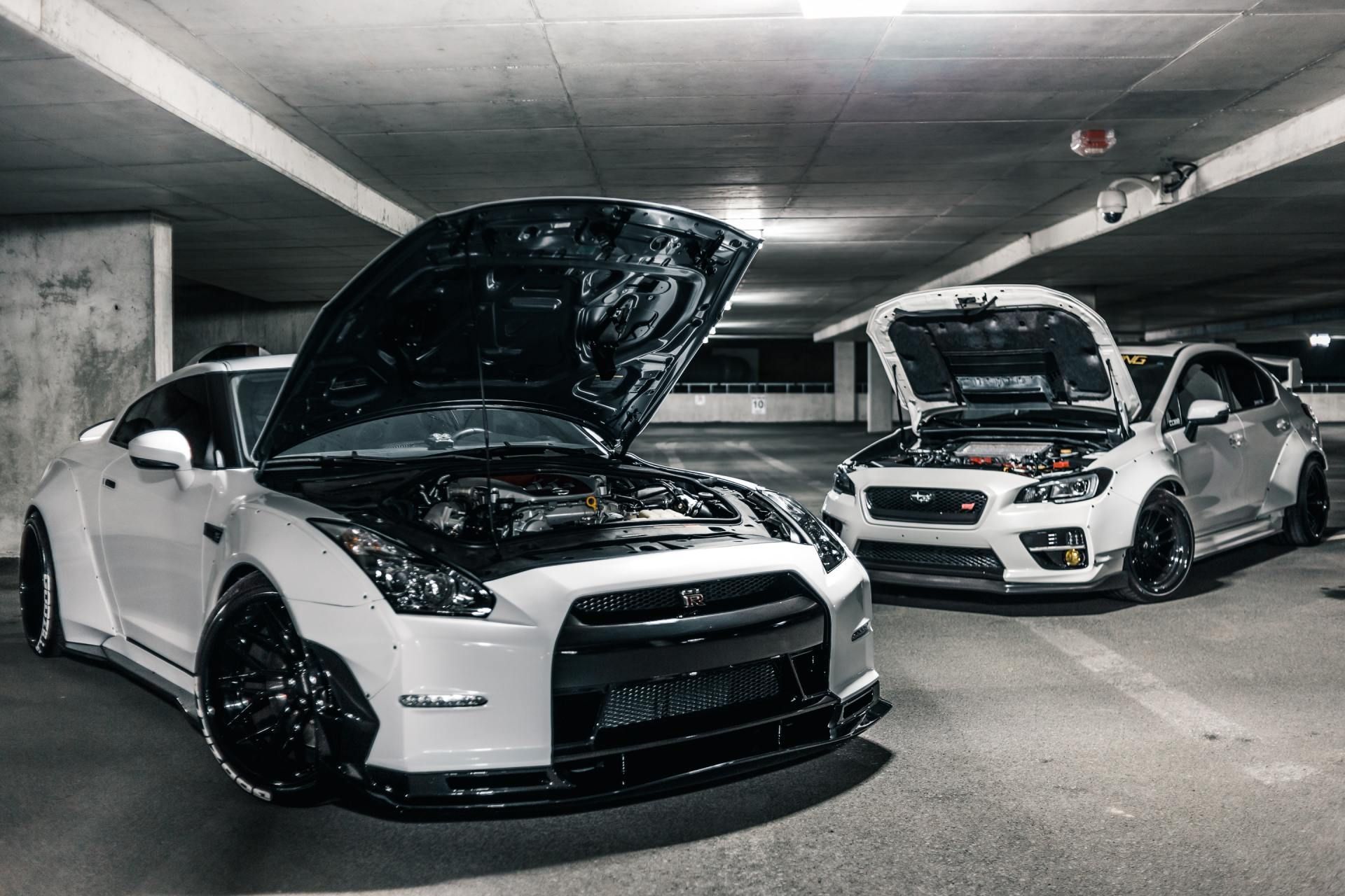 White Nissan GT-R and Subaru WRX with hoods open, parked in a dimly lit parking garage.