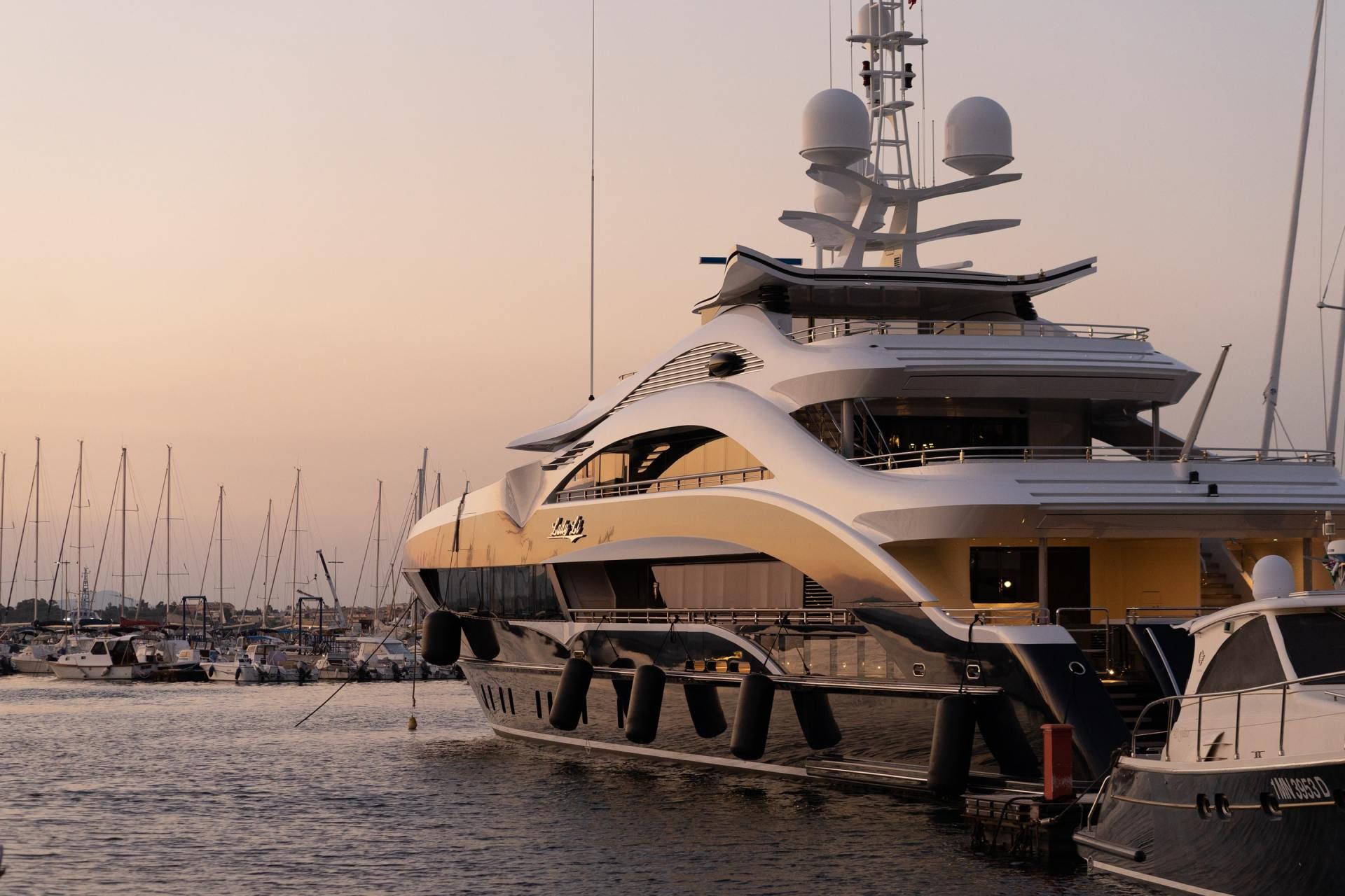 Luxury yacht docked at a harbor at sunset. Gold and black hull, white superstructure, masts with radar.