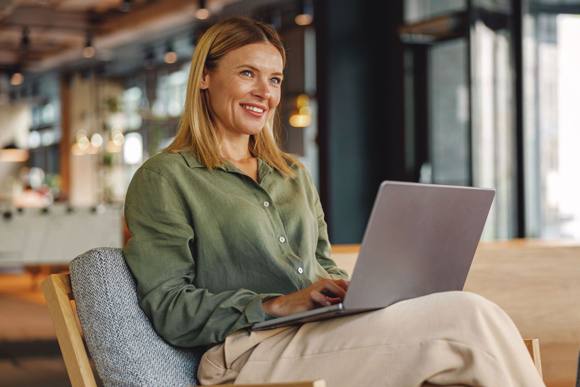 Woman with blonde hair smiling while using a laptop, seated indoors in a modern setting.