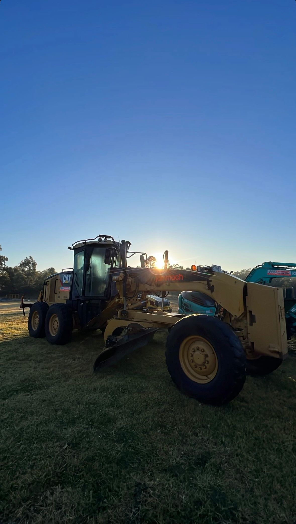 Yellow CAT grader parked on grass at sunset, with construction equipment in the background. — Jammach Earthmoving in Newcastle, NSW