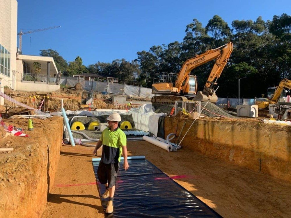 A Man is Walking Through a Construction Site With an Excavator in the Background — Jammach Earthmoving in Central Coast, NSW
